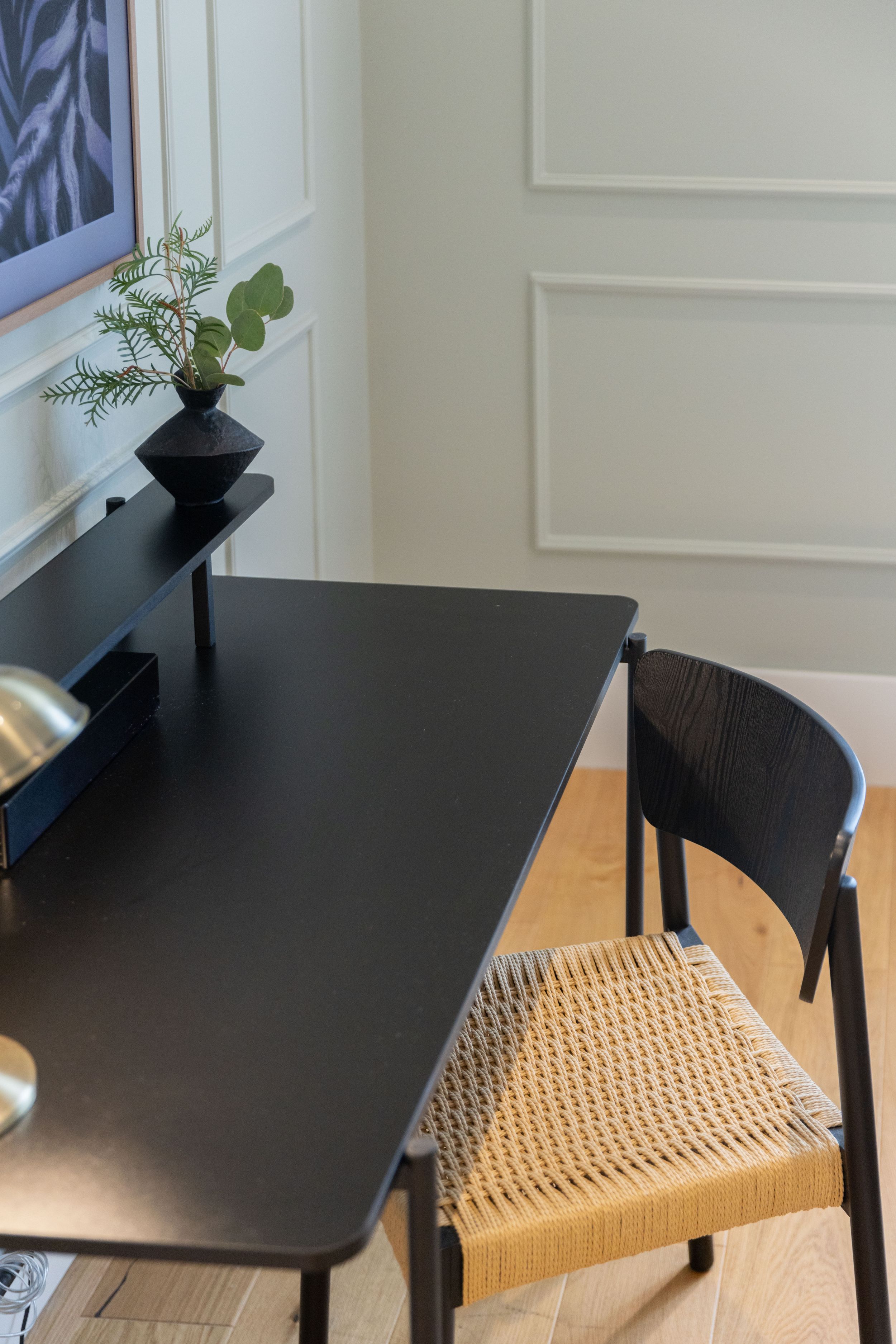 Modern black desk with woven chair seat and small plant in geometric vase against white paneled wall.