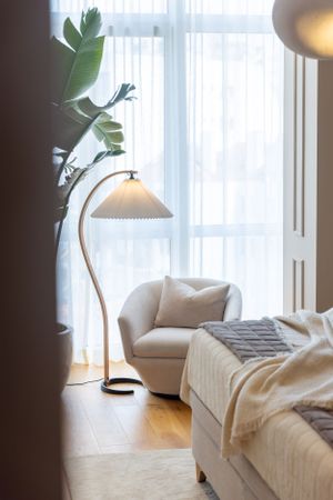 Cozy reading nook with curved floor lamp, white armchair, and large indoor plant by sheer curtains.