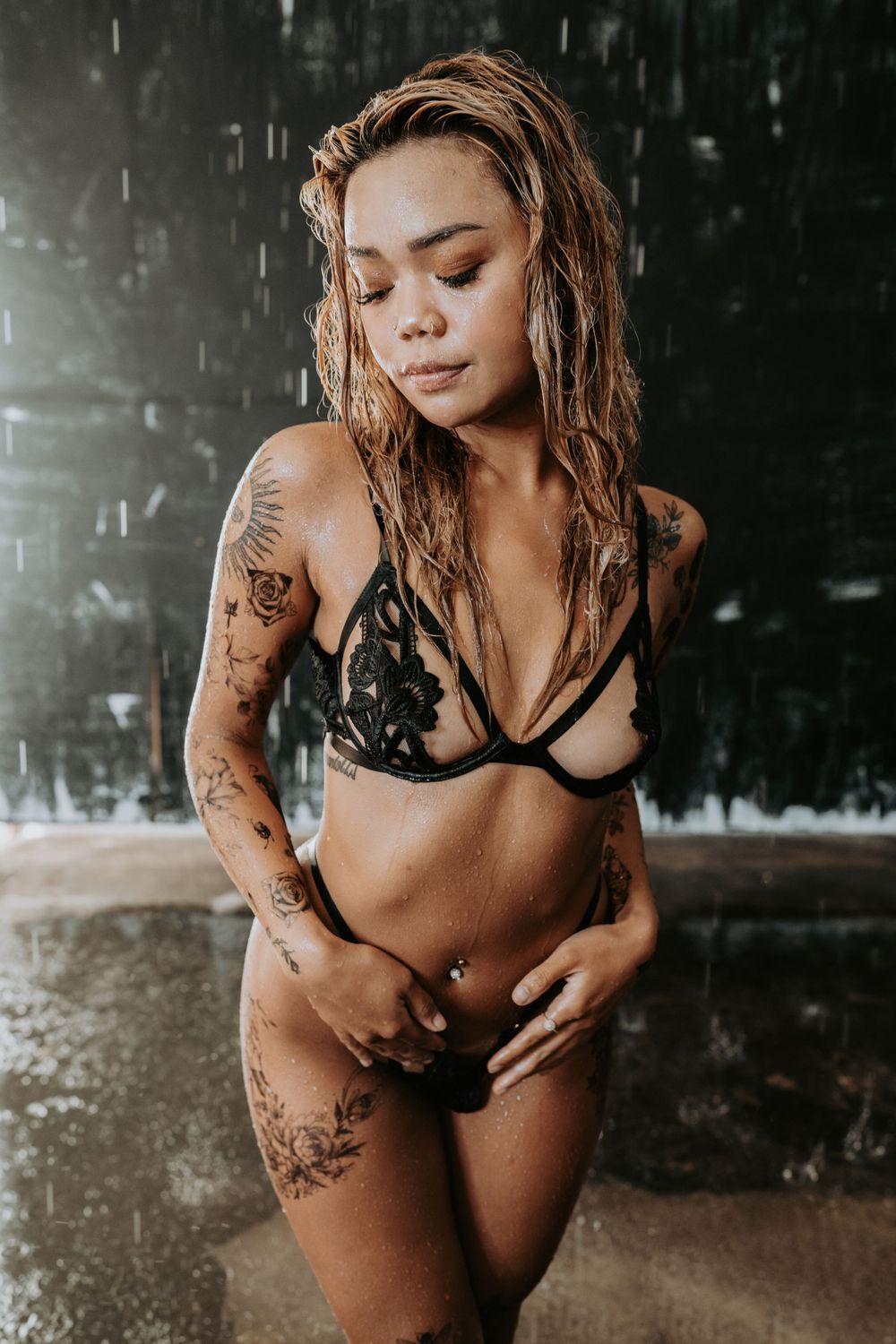 Tattooed model in black lingerie poses in artistic rain photography setting.