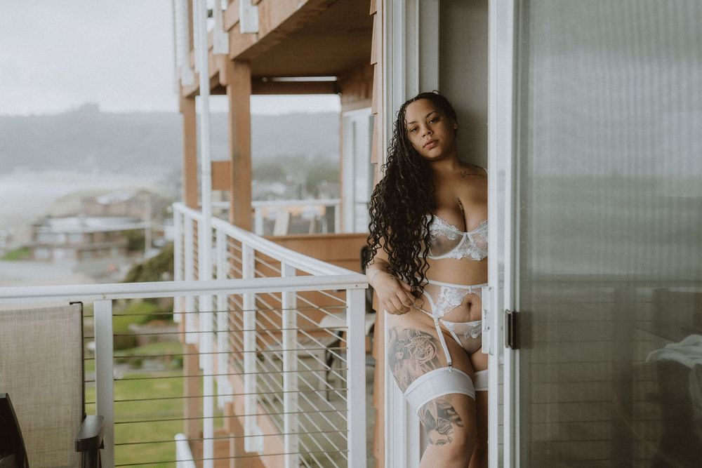 Woman in white lingerie posing on a balcony with ocean view, featuring curly hair and thigh tattoo.