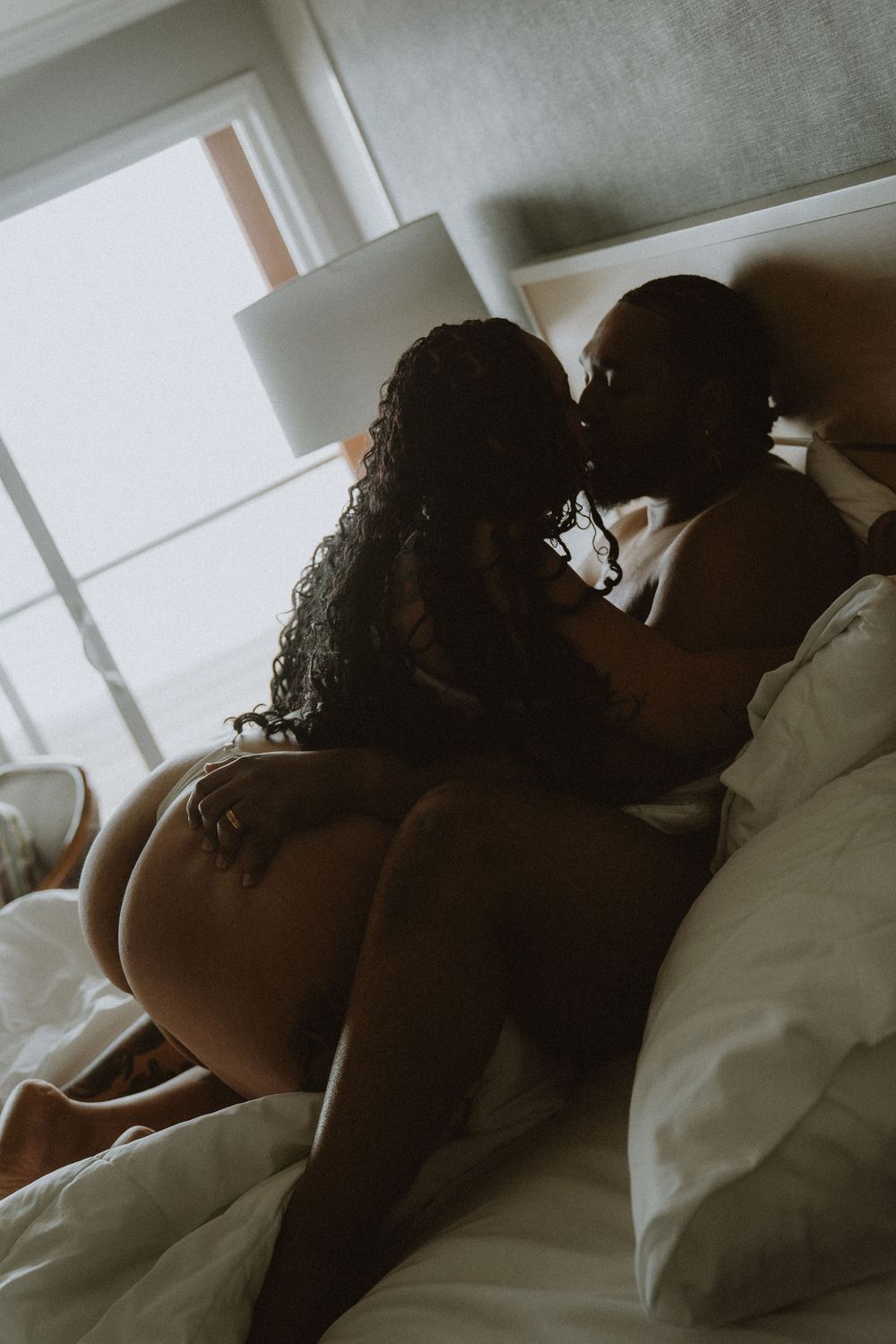 Silhouette of a couple kissing intimately on a white bed in a softly lit bedroom.