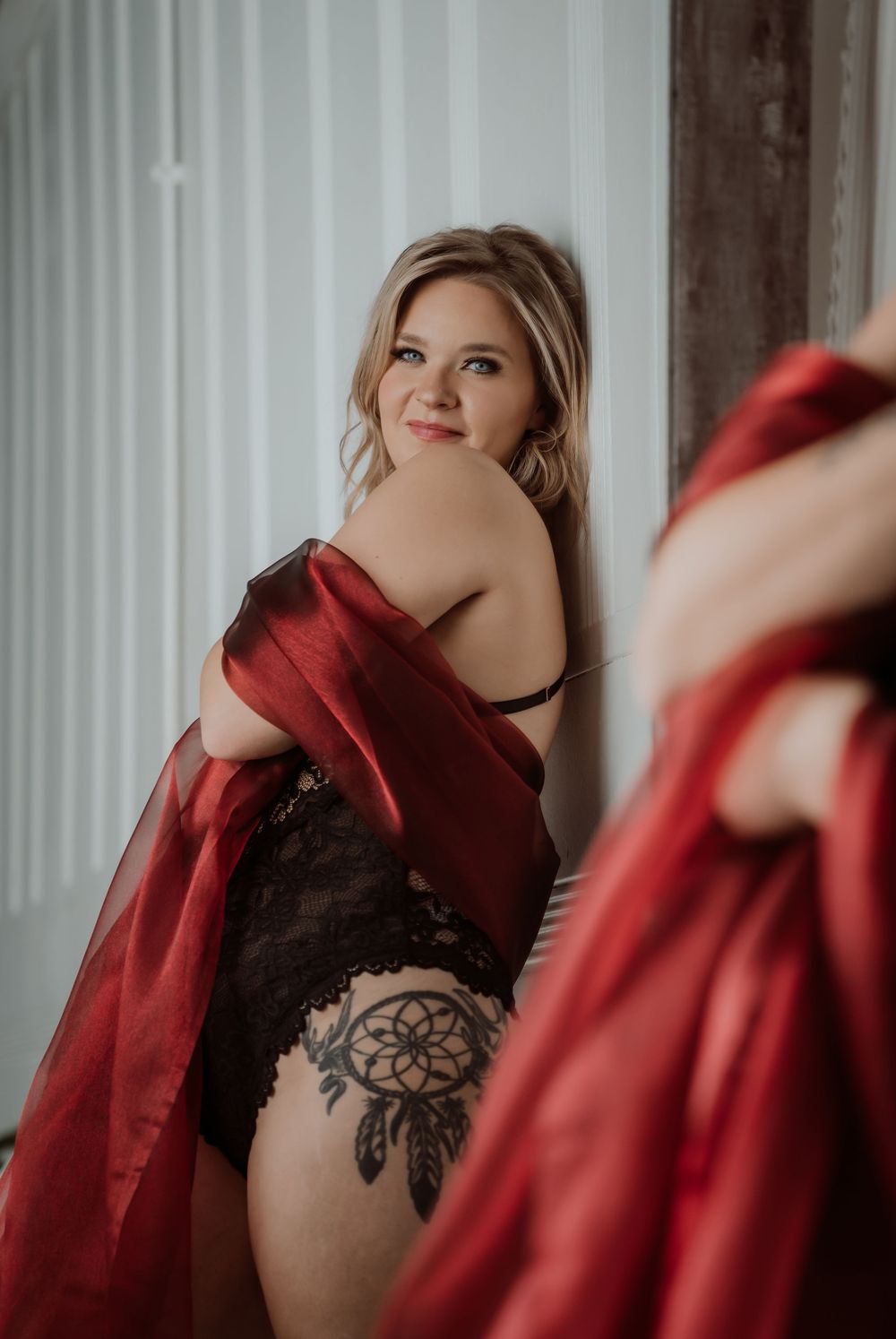 Woman in black lace bodysuit and red silk robe with dreamcatcher tattoo poses elegantly in boudoir photography.