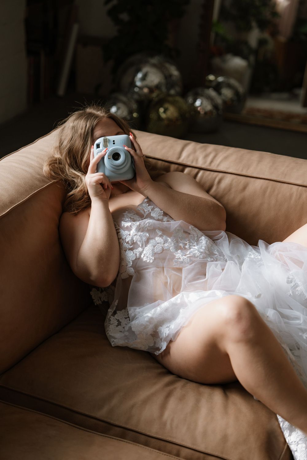 Bride in white lace lingerie lying on a couch taking a photo with a blue Instax camera.