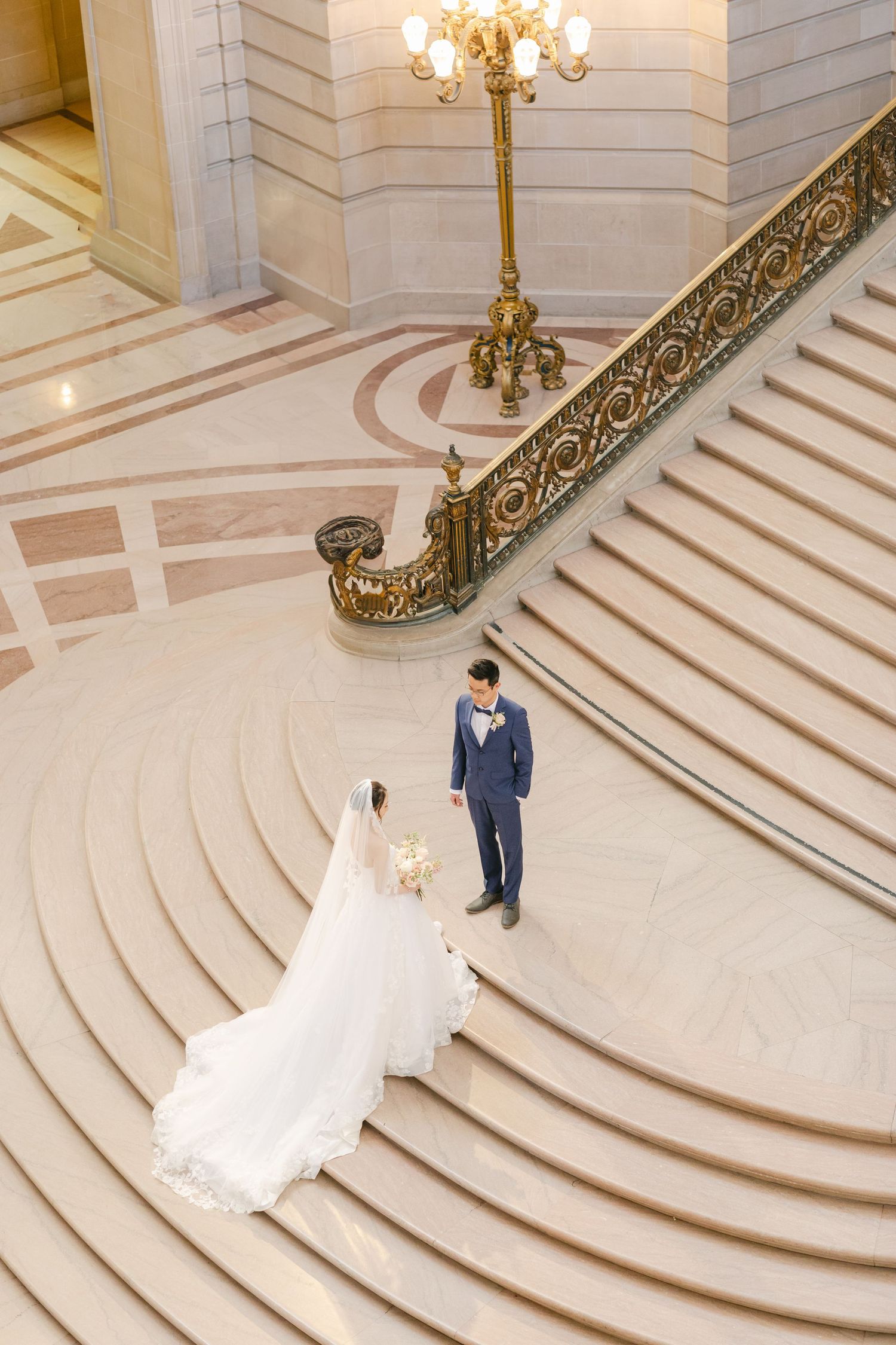Elegant wedding couple standing on grand marble staircase with ornate gold railing and chandelier in luxury venue.By Chelsee Rawe Photograph