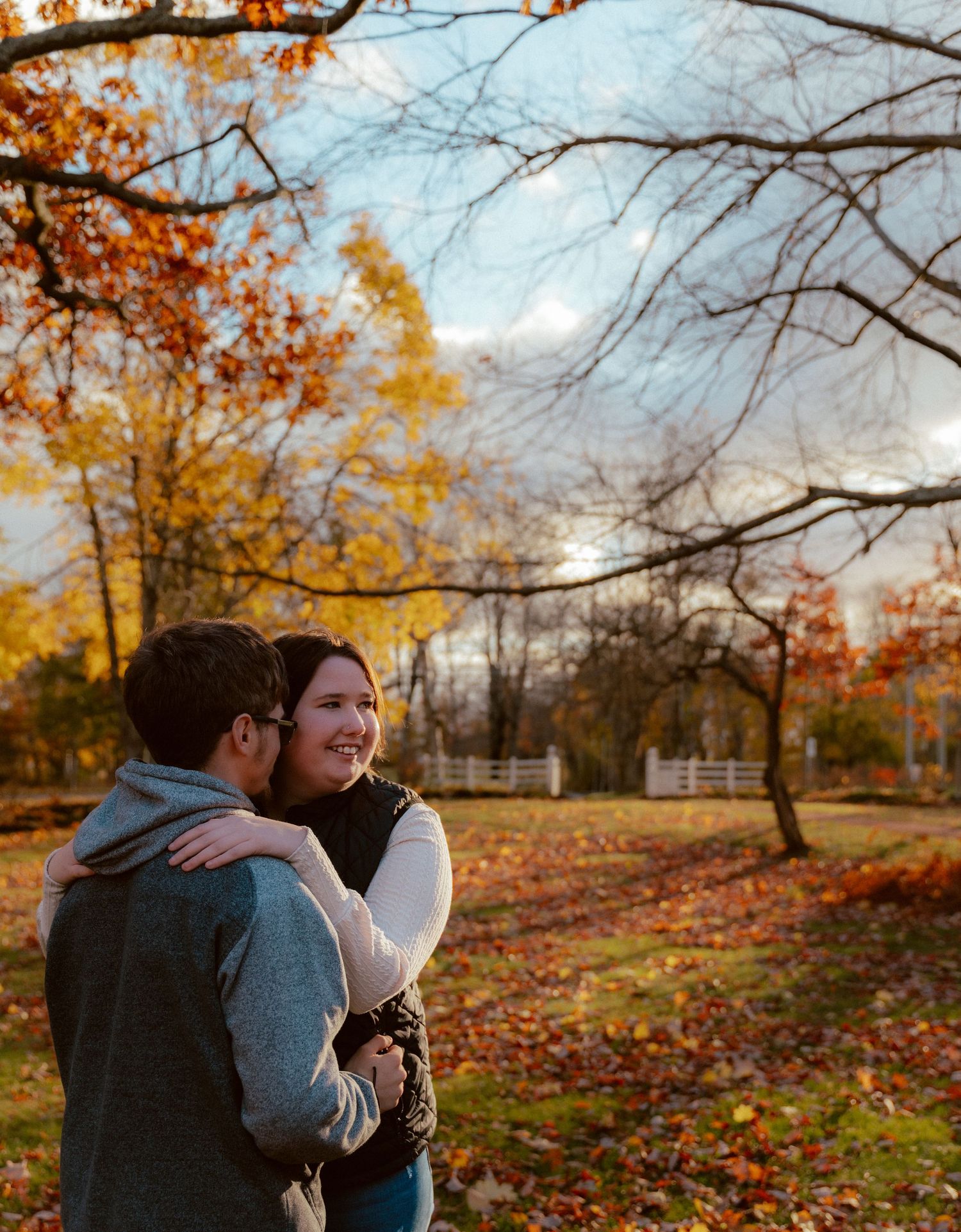 Couple embracing in autumn park with golden leaves and white fencing in background.