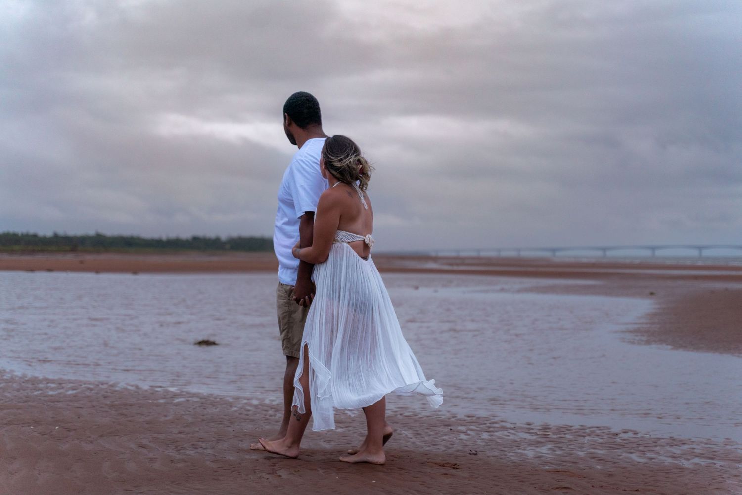 A couple in white clothing stands together on a misty beach at dusk.