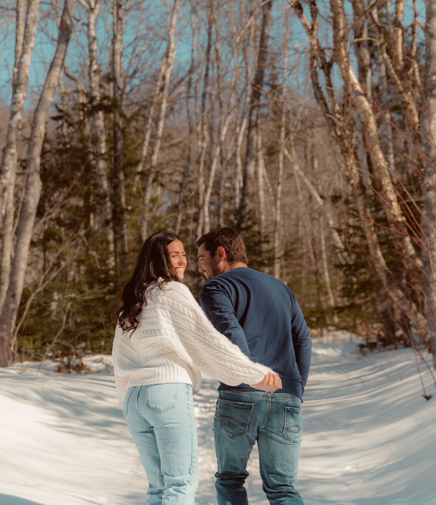 A couple walking hand in hand on a snowy forest path surrounded by bare winter trees.