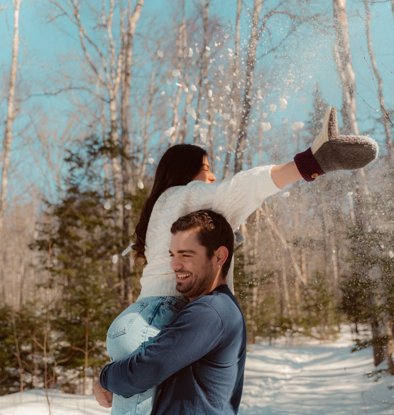 A couple plays together in a snowy winter forest setting with bare trees in the background.