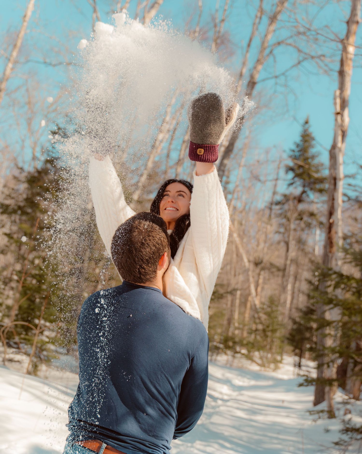 A playful winter scene captures someone being lifted up while throwing snow in the air against a backdrop of bare trees.
