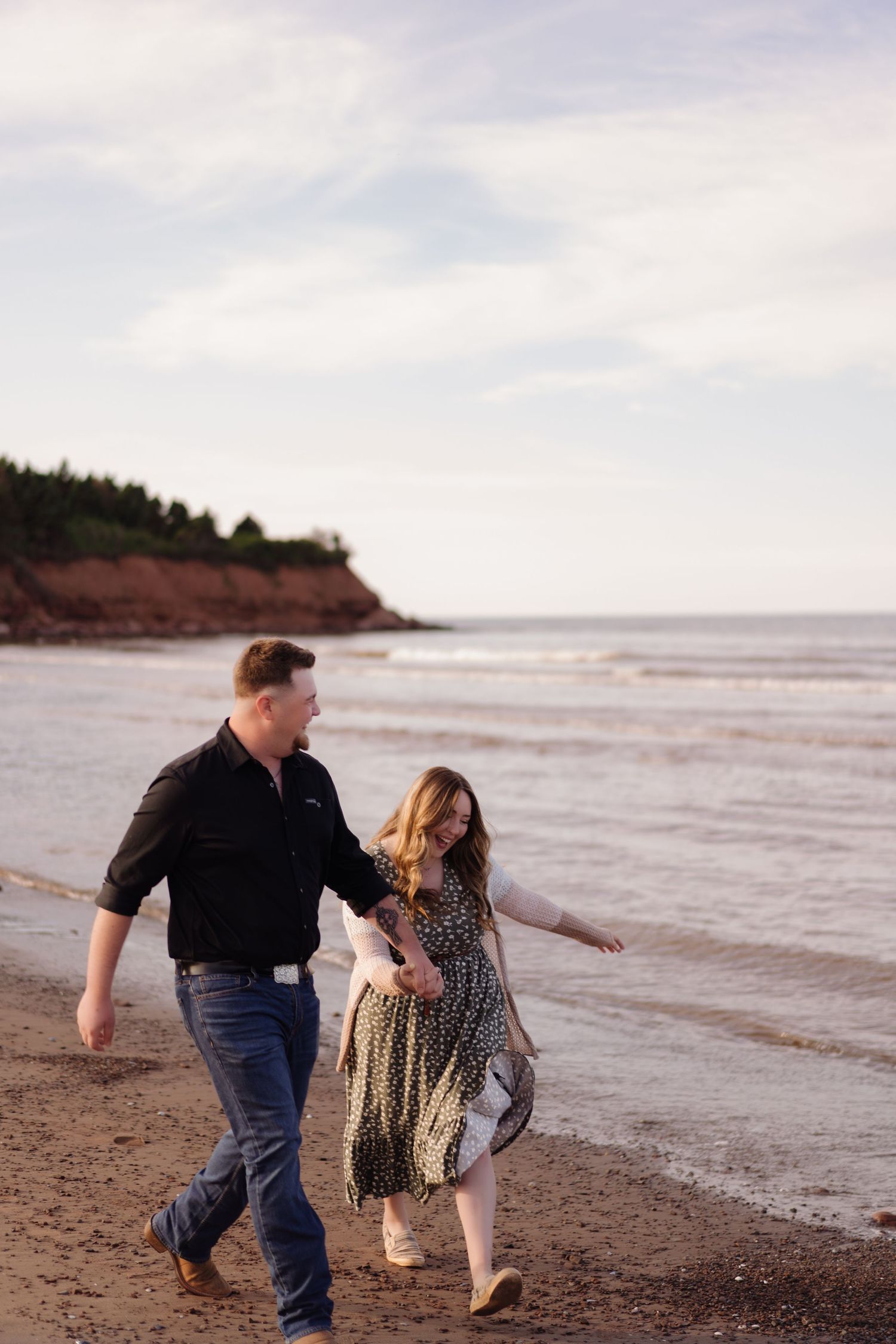 A couple walks playfully along a scenic coastal beach at sunset with water and cliffs in the background.