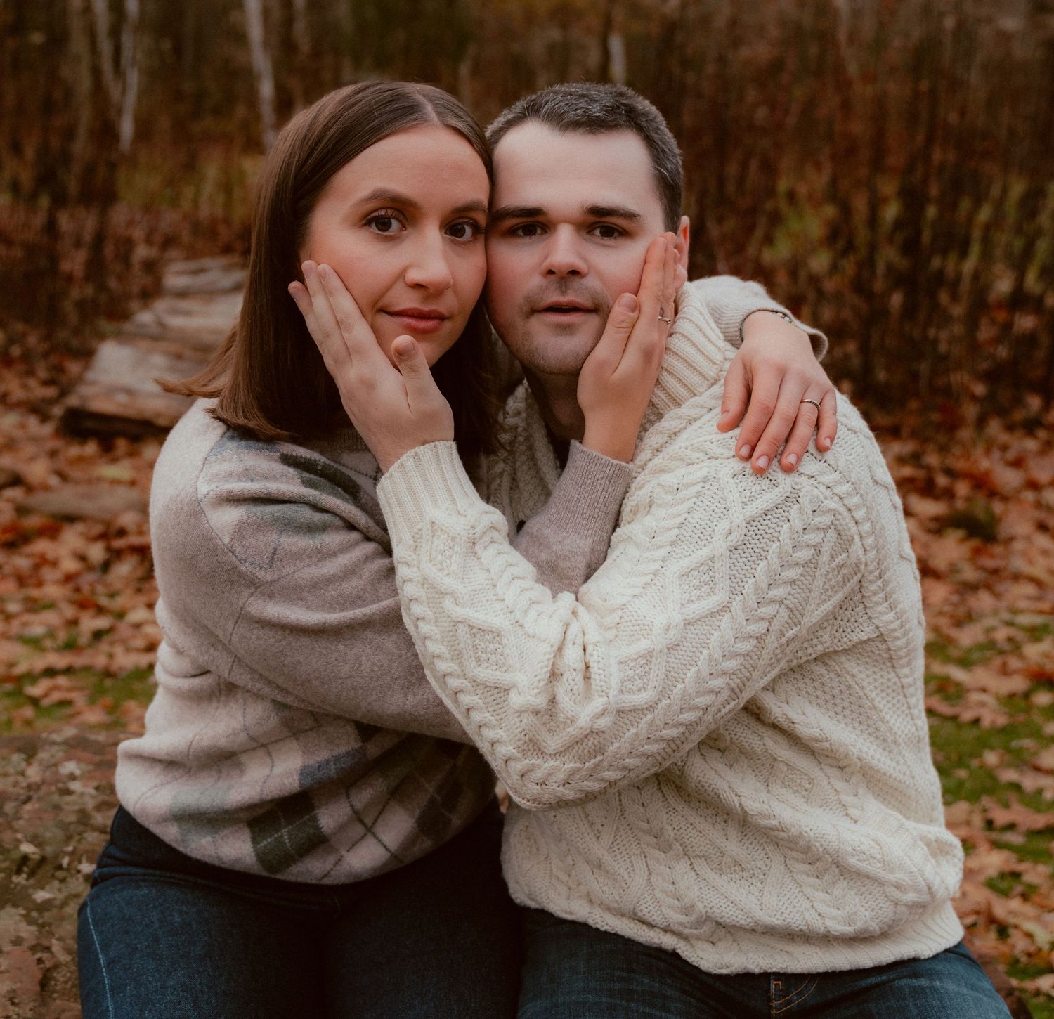 A couple in cozy sweaters embrace while posing on an autumn path surrounded by fallen leaves.
