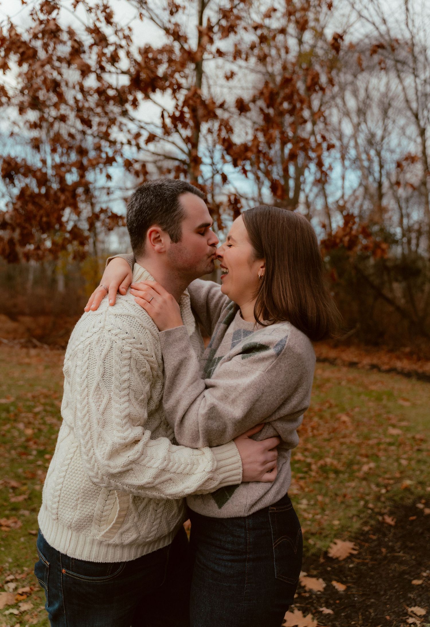 A romantic autumn couple embrace wearing cozy sweaters among fall foliage and trees.
