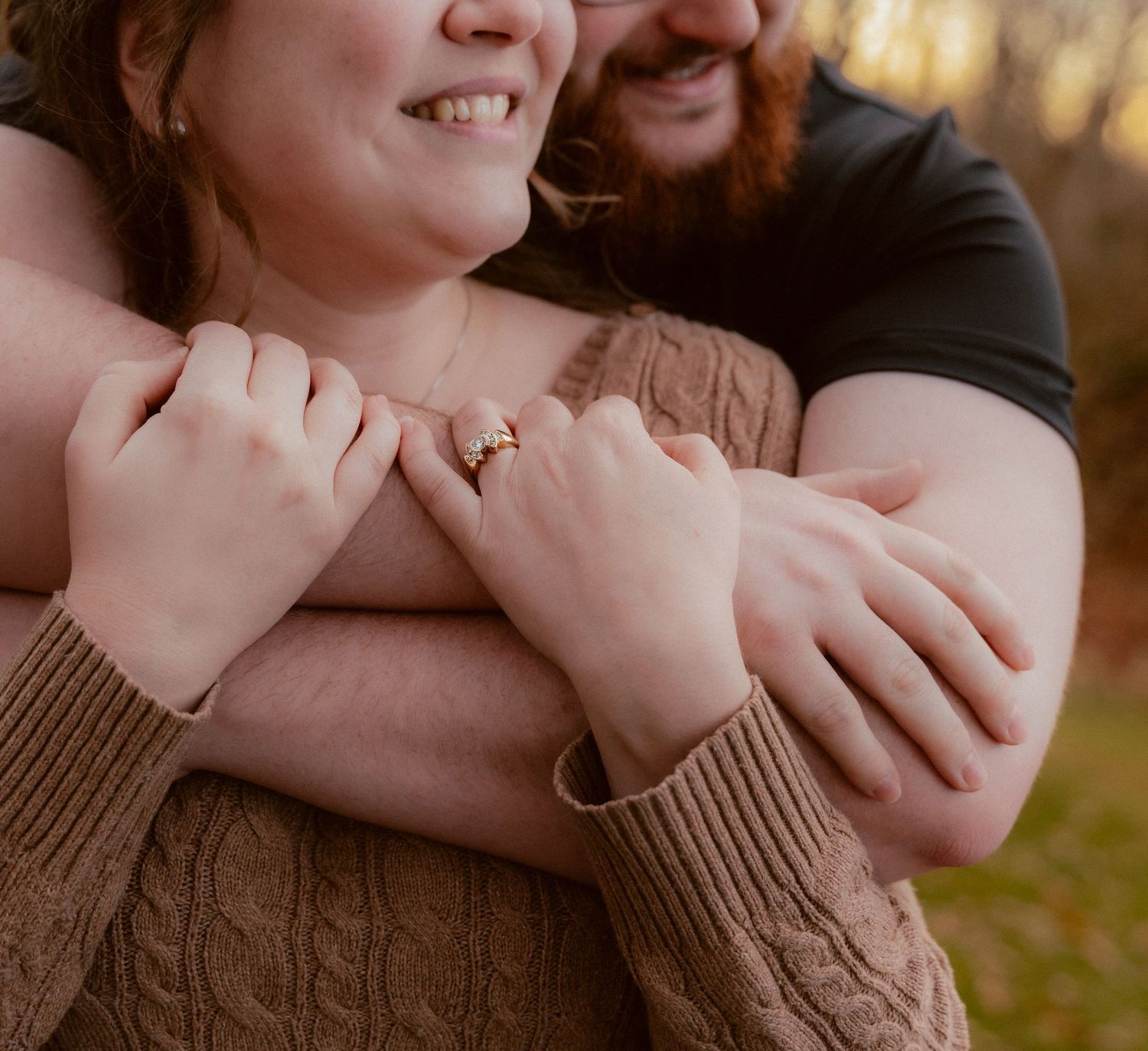 Close-up of embracing couple showing engagement ring on her hand with brown sweater sleeve and outdoor setting.