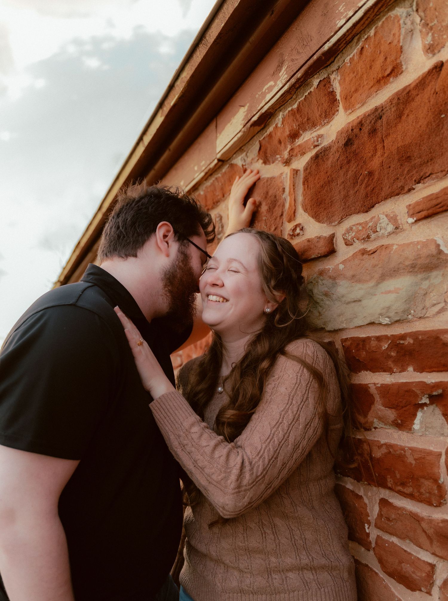 A couple shares an intimate moment leaning against a rustic brick wall while exchanging joyful smiles and laughter.