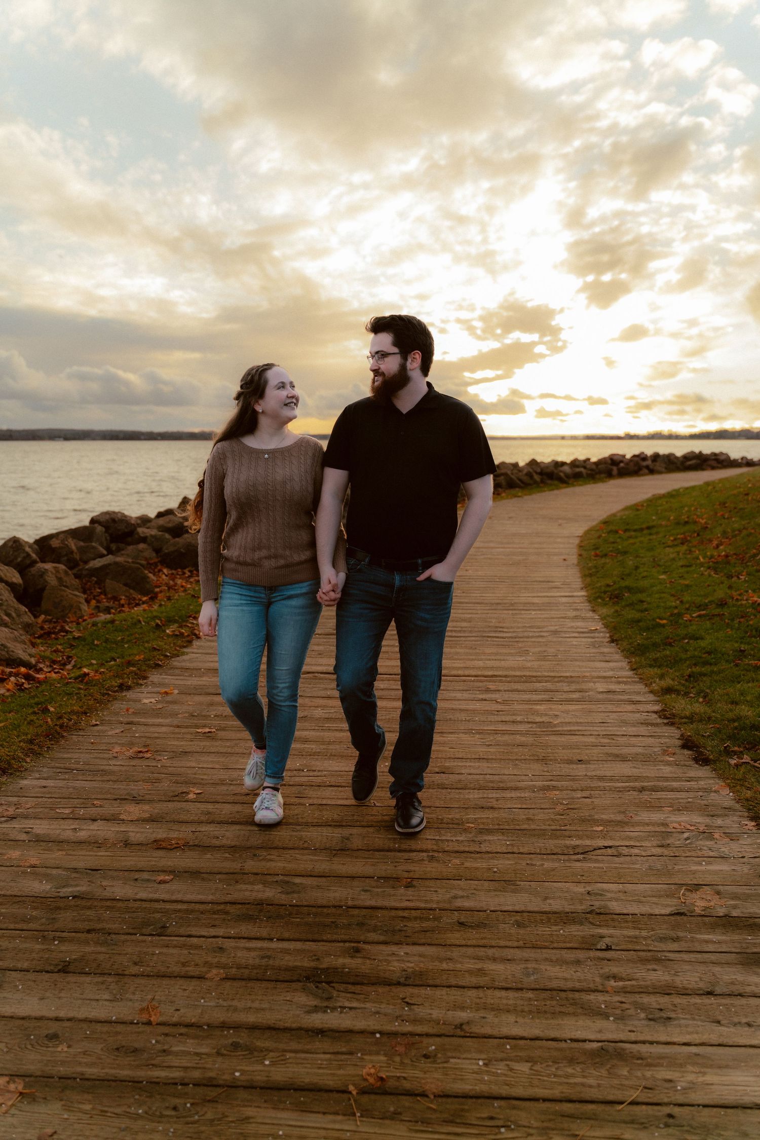 A couple walks together on a wooden boardwalk by the water during a scenic sunset with dramatic clouds.