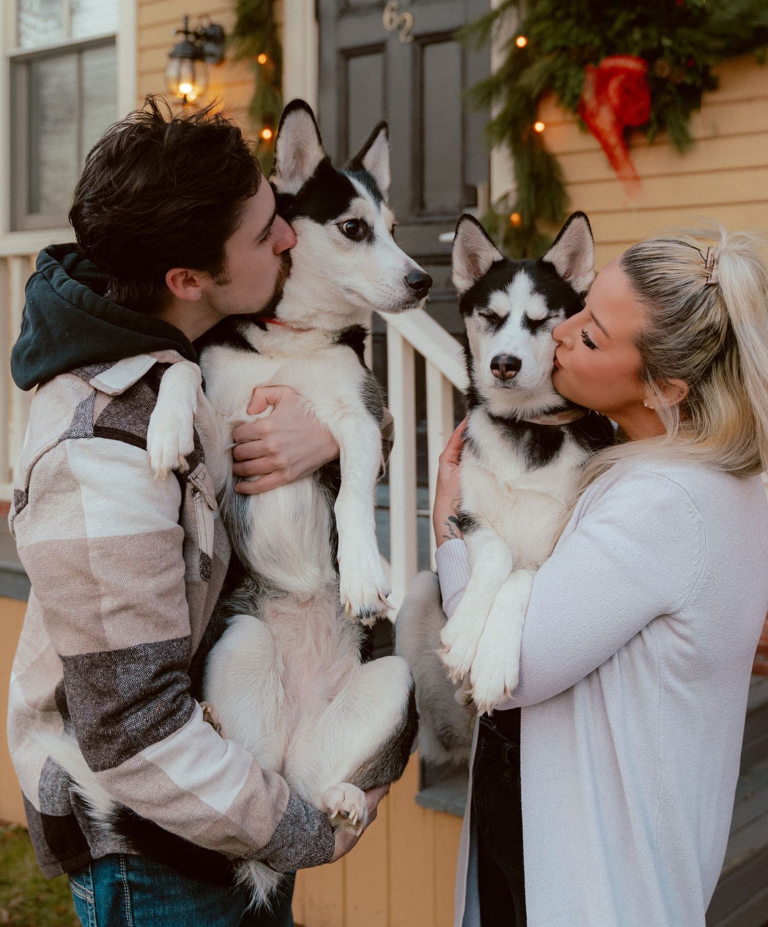Couple sharing kisses with their two Husky dogs on decorated holiday porch.