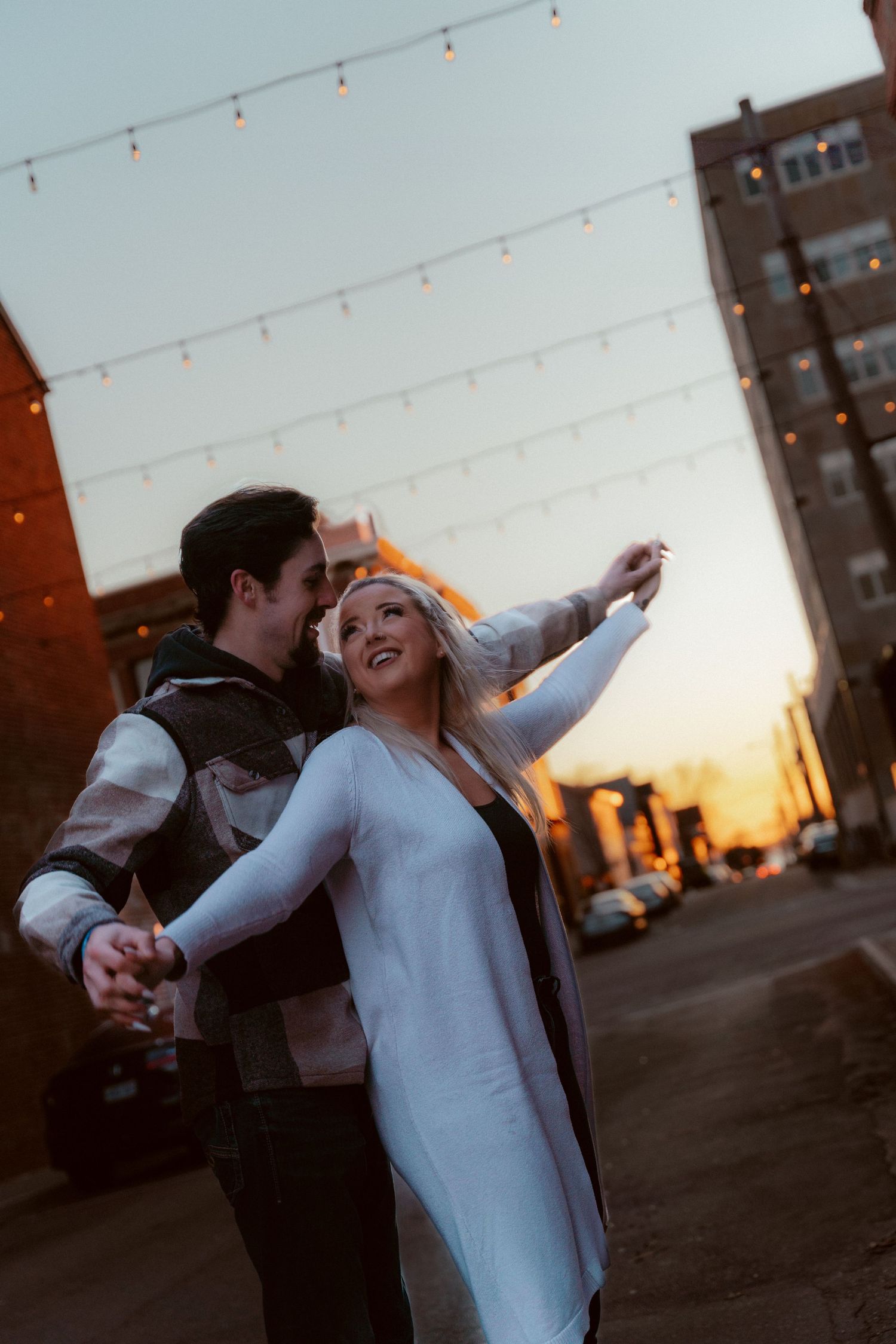 Dancing couple laughing together at sunset with string lights overhead in urban setting.