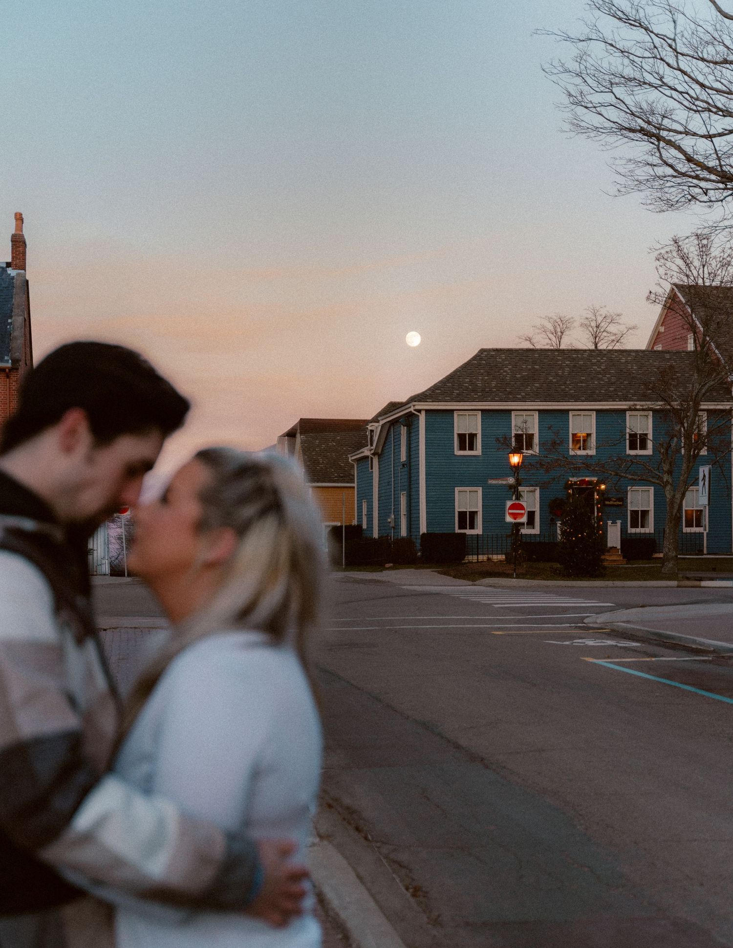 A couple embraces at dusk in front of a blue house as the moon rises in the evening sky.