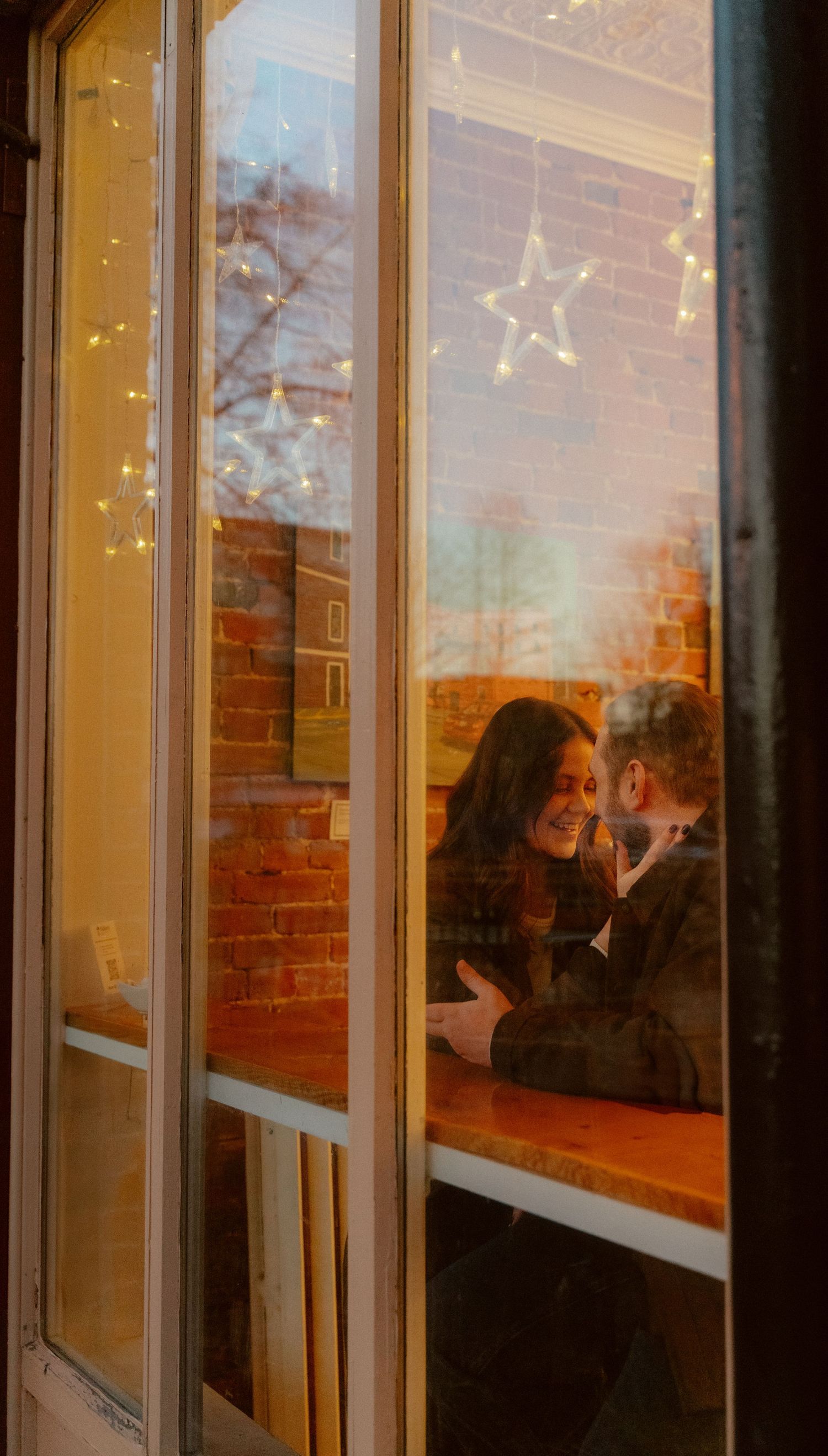 A couple sits by a window decorated with glowing star lights in a romantic evening setting.
