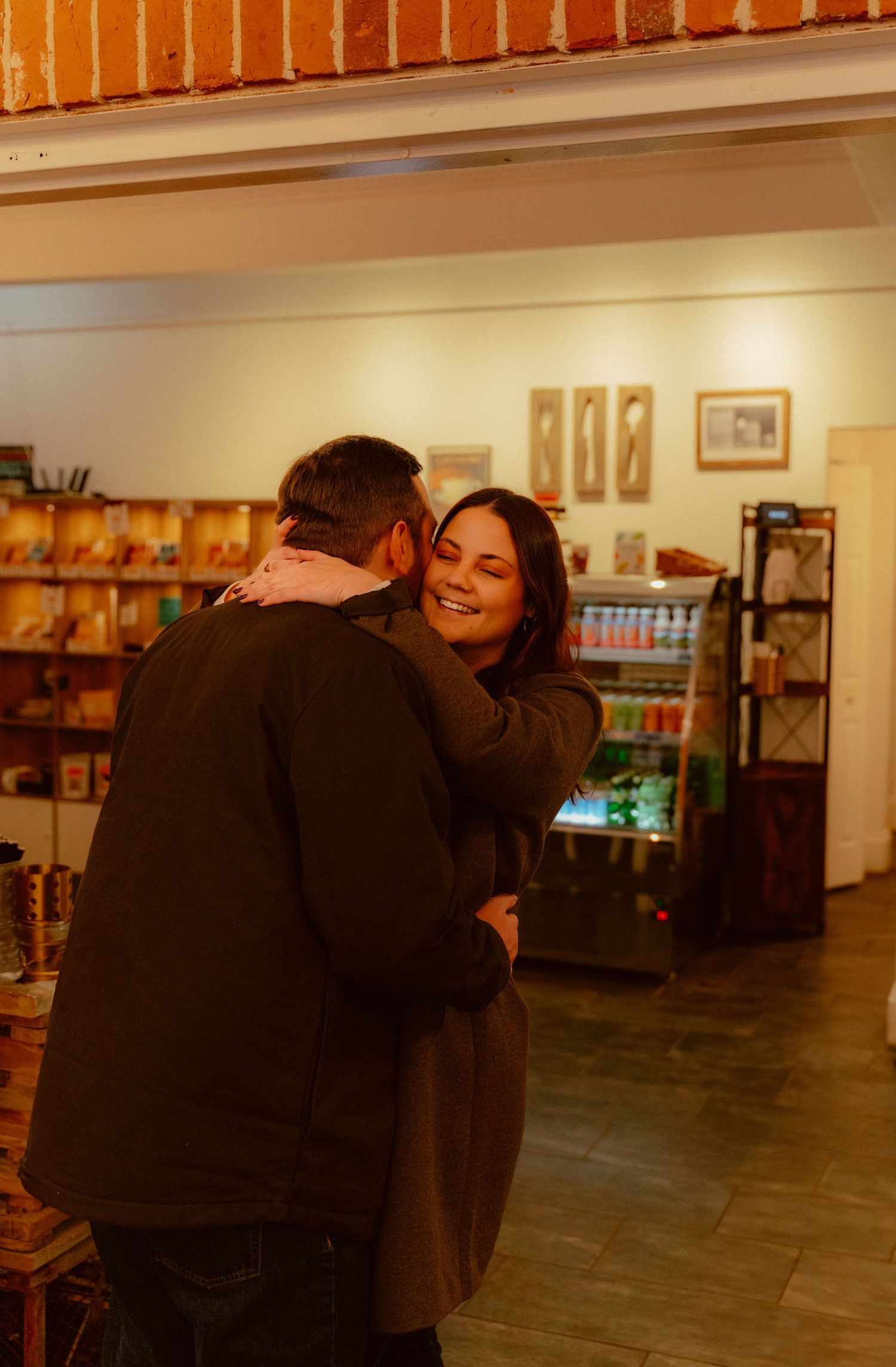A couple shares a warm embrace while standing in a restaurant with wooden floors and shelves visible in the background.