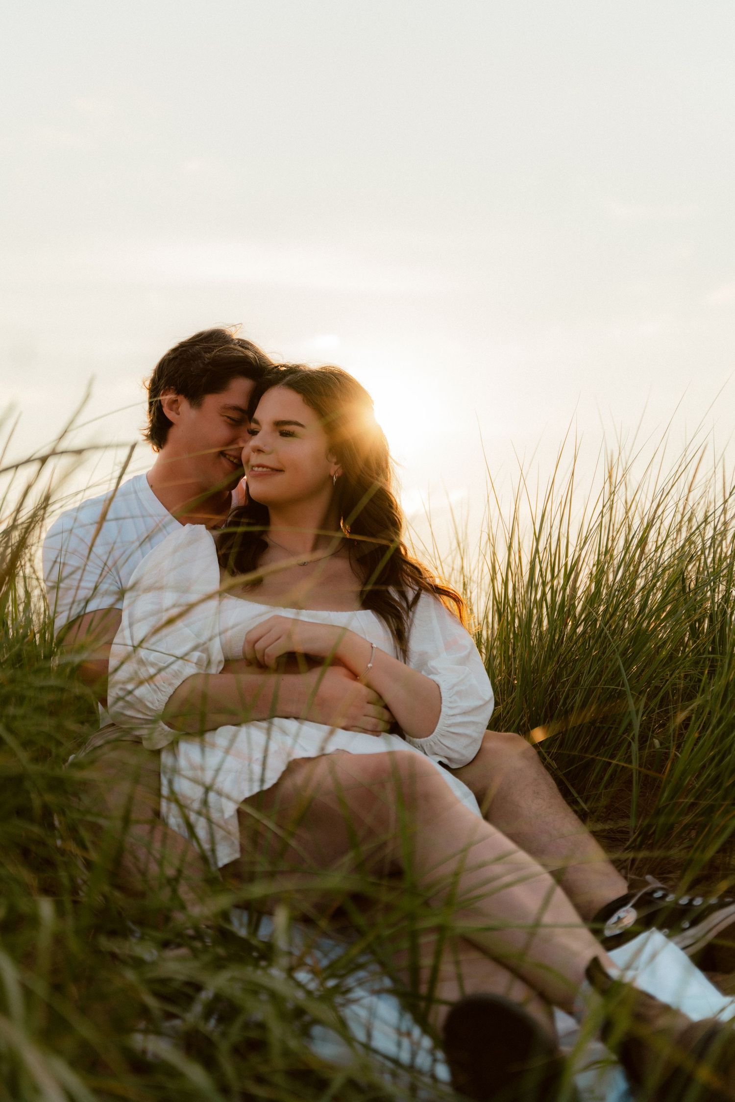 A tender sunset moment of a couple sitting in tall grass by the ocean.
