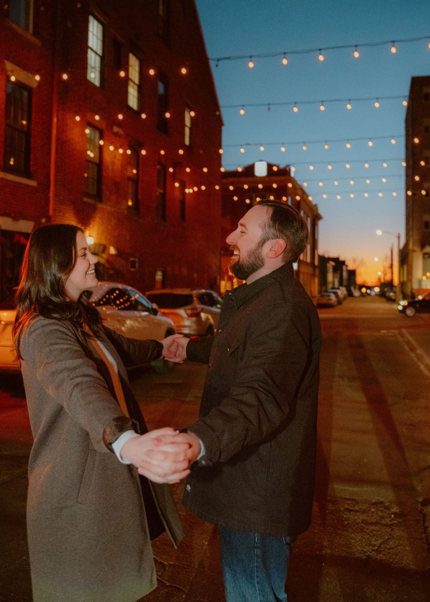 A couple shares a romantic dance moment on a city street at dusk under strung lights and brick buildings.