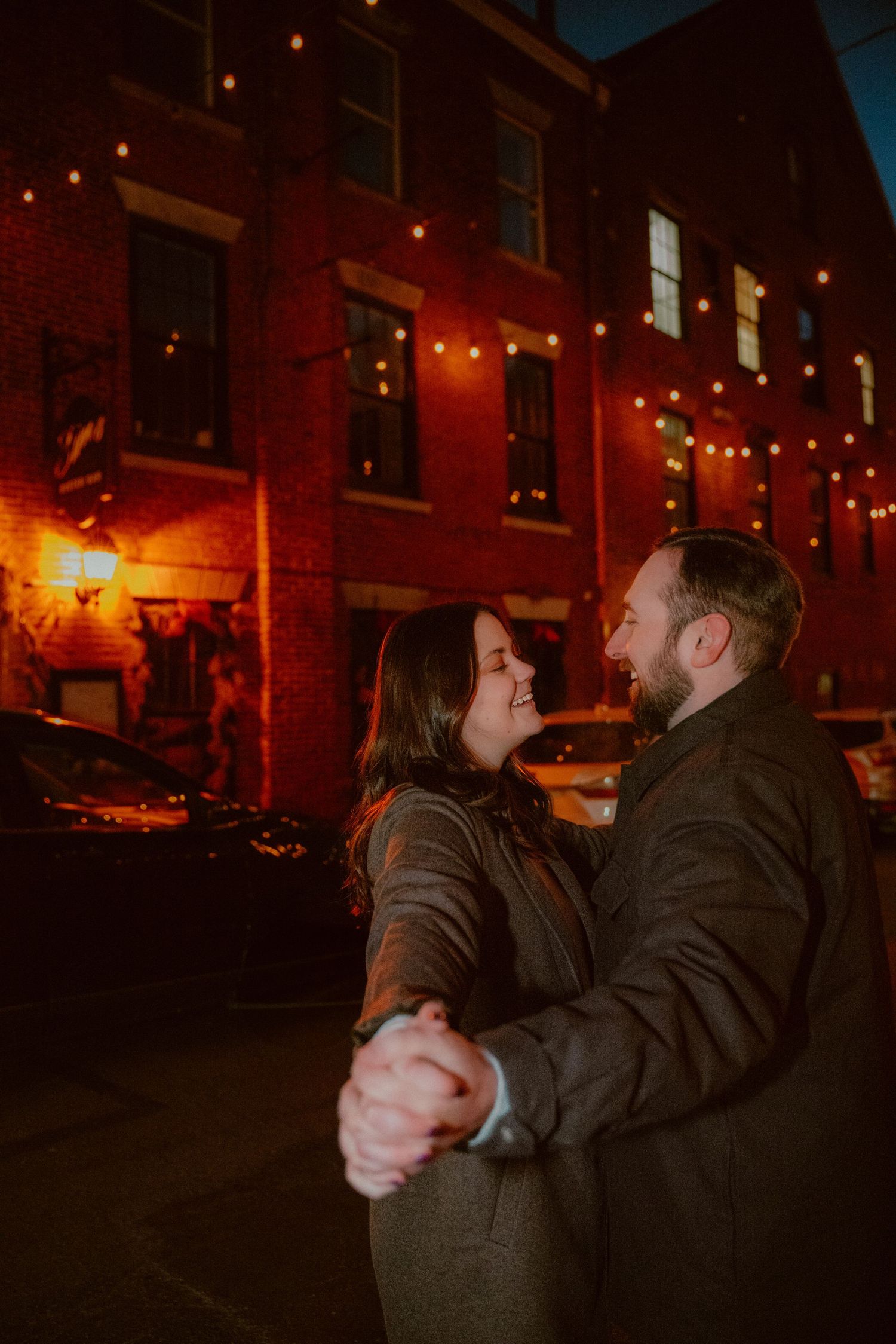 A couple dances together at night under warm string lights by a brick building on a city street.