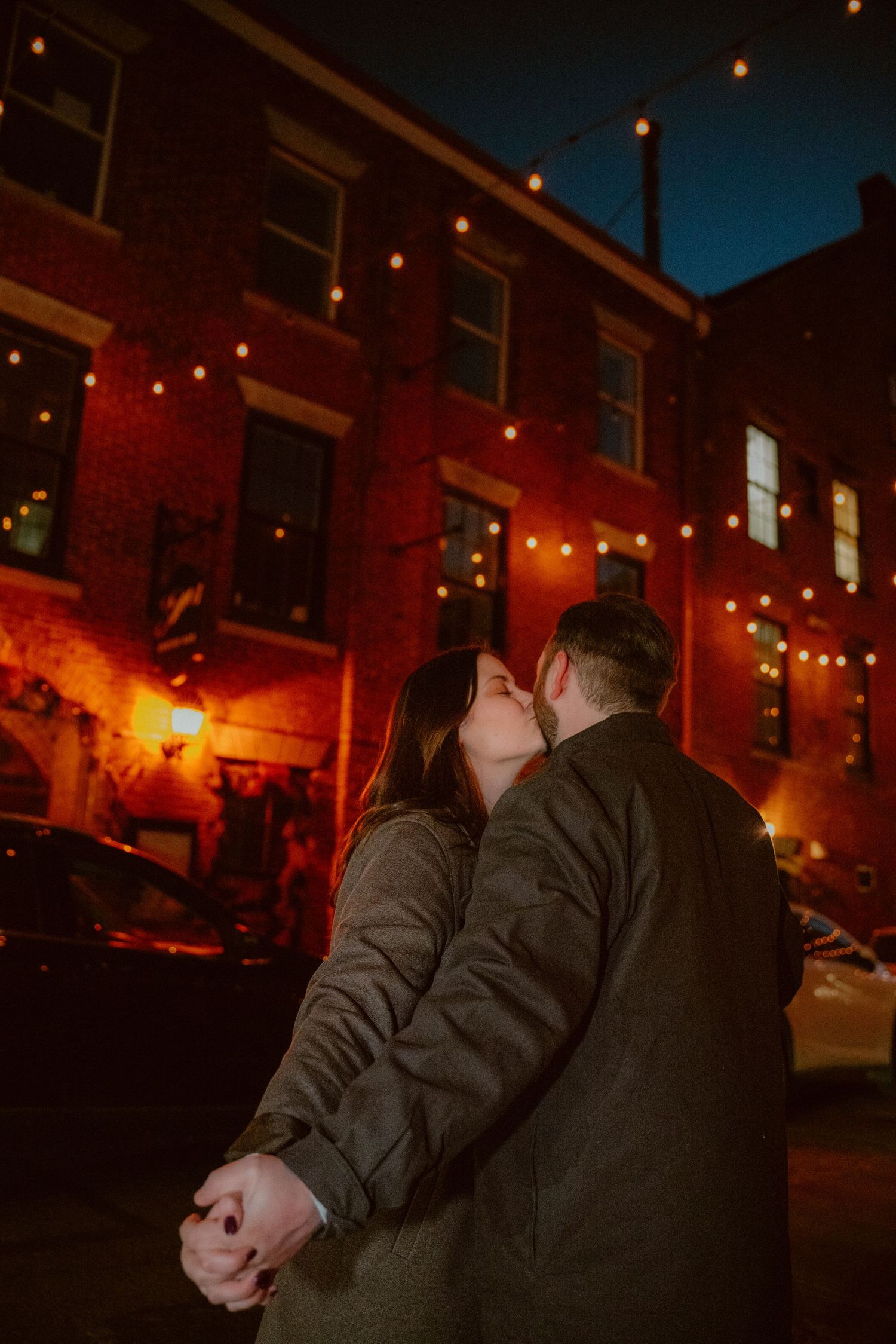 A romantic couple shares a kiss under glowing string lights against a red brick building on a dark evening.