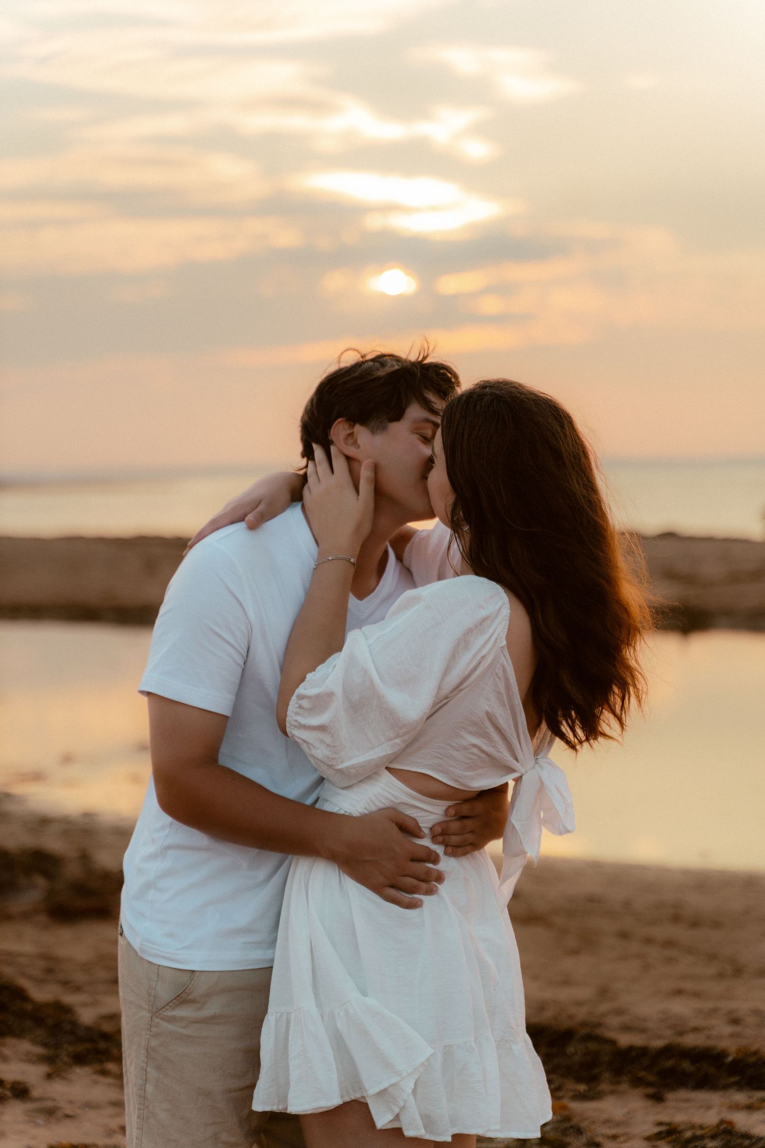Couple kissing on beach at sunset with golden light reflecting off water.