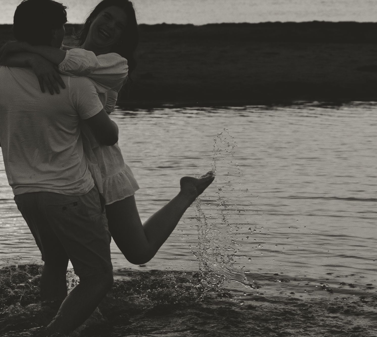 A romantic black and white photo of a couple embracing by a tranquil lake at sunset.