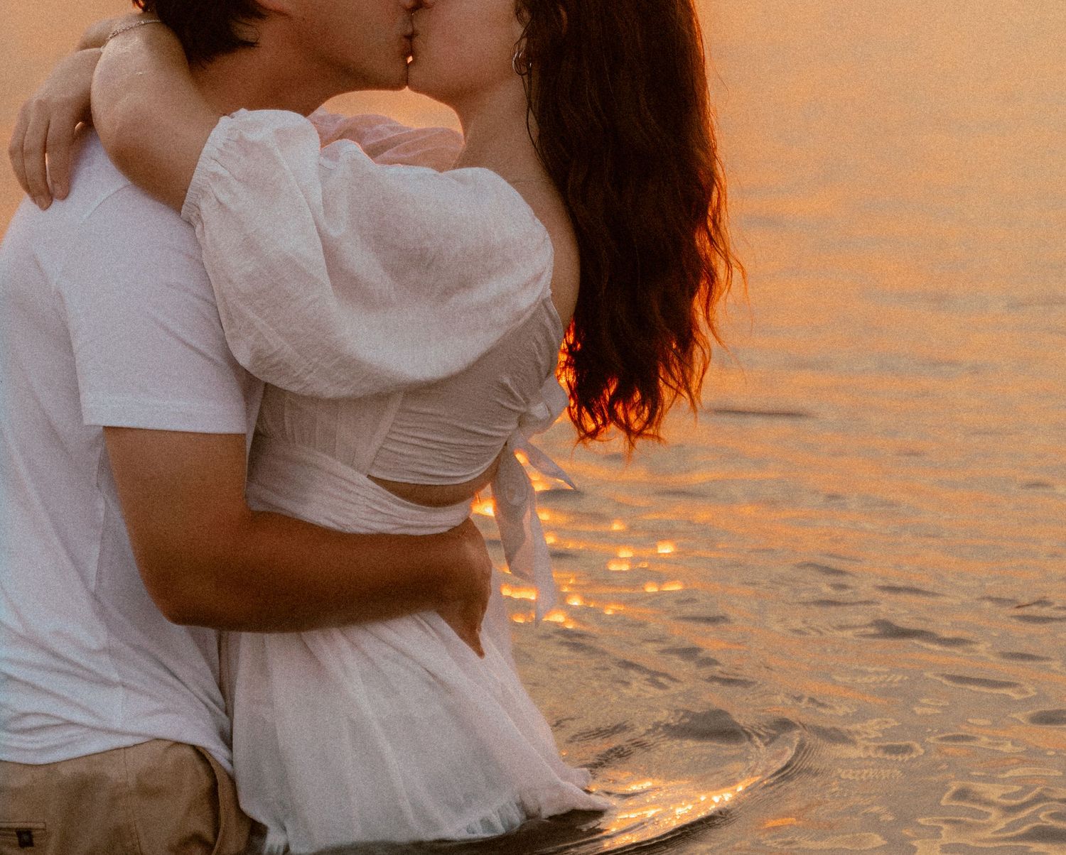 A couple in white clothing sharing an intimate moment by the water during golden hour.