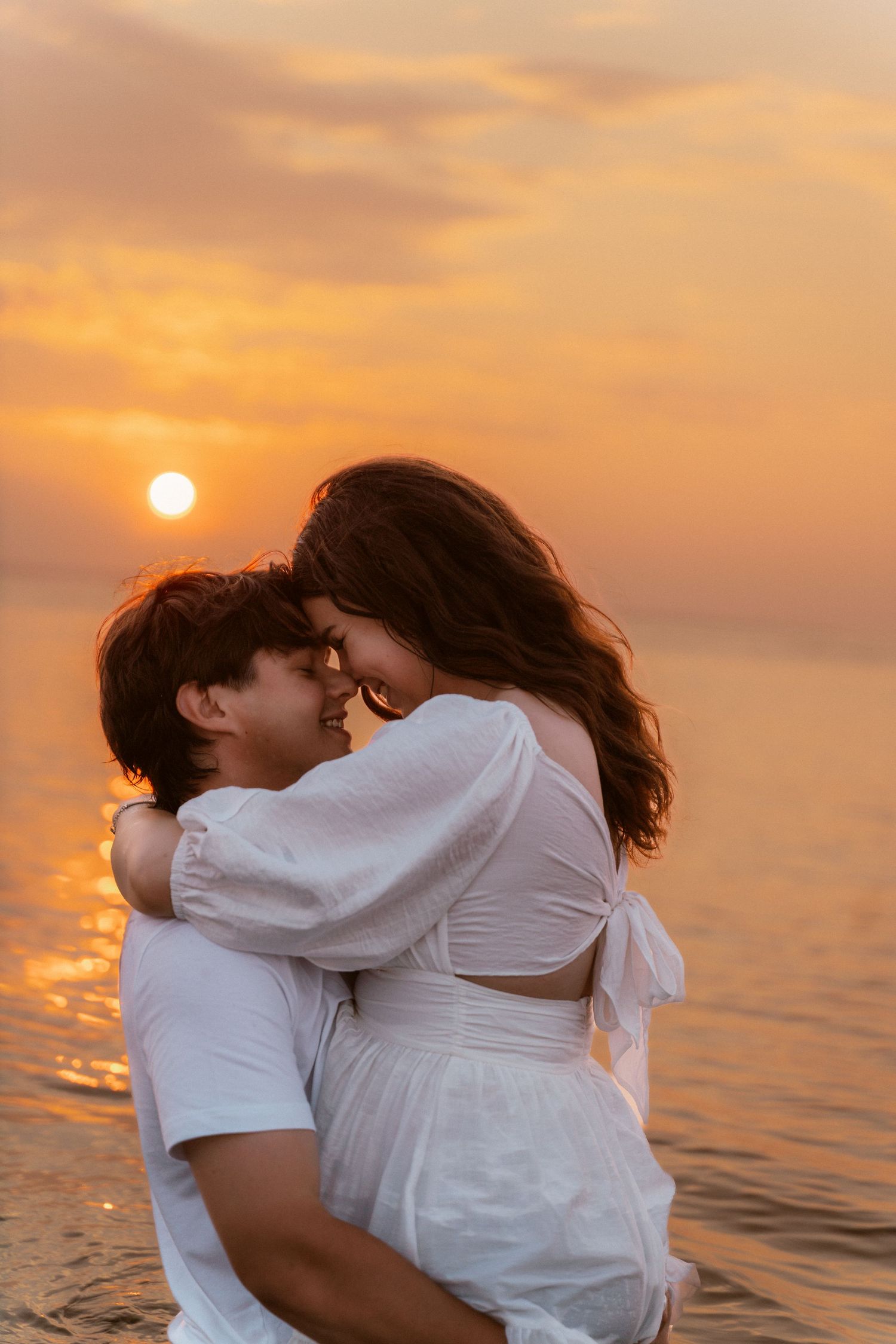 A romantic couple in white clothes embracing against a vibrant orange sunset over the ocean.