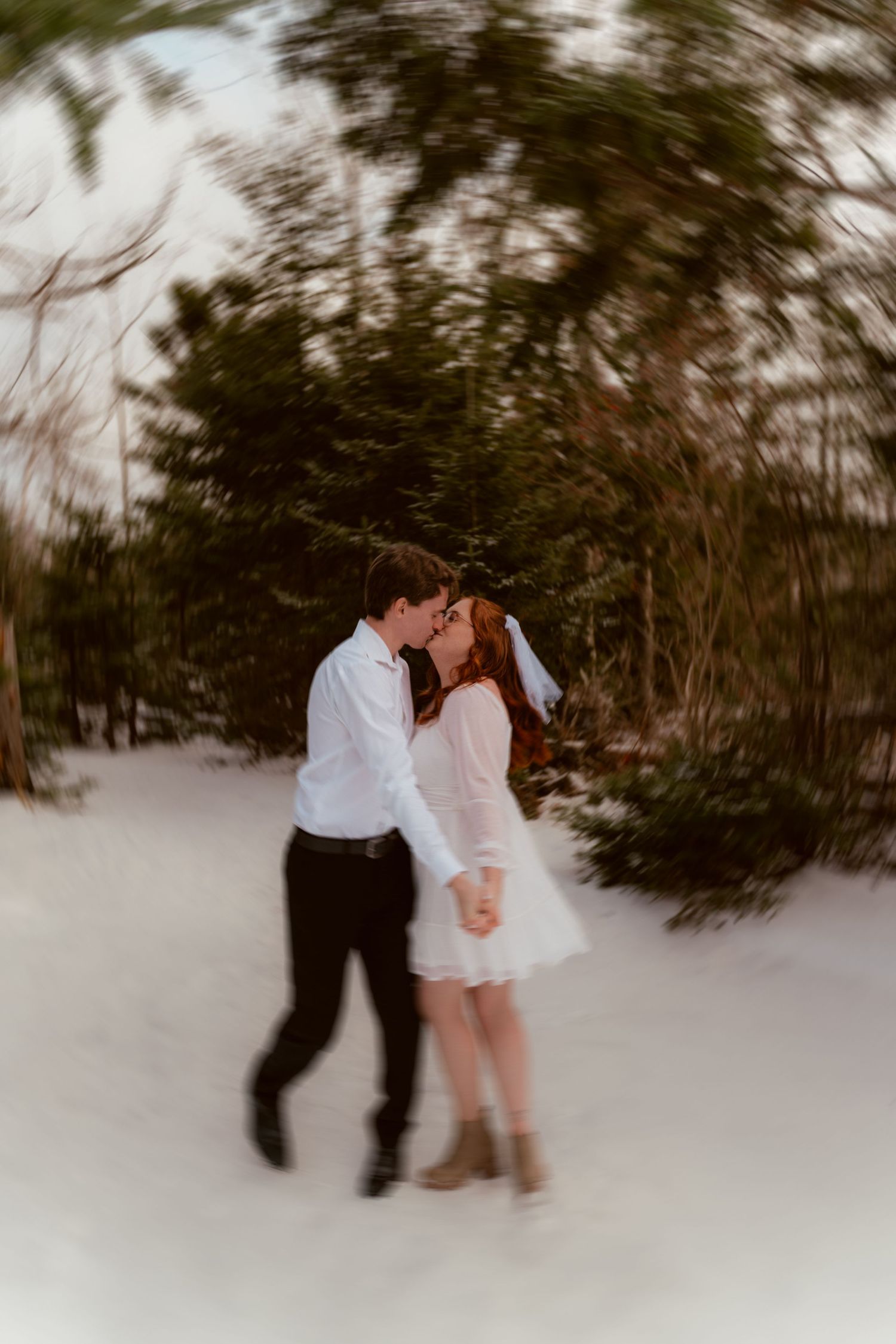 A romantic winter engagement photo of a couple in white sharing an intimate moment in the snow-covered forest.