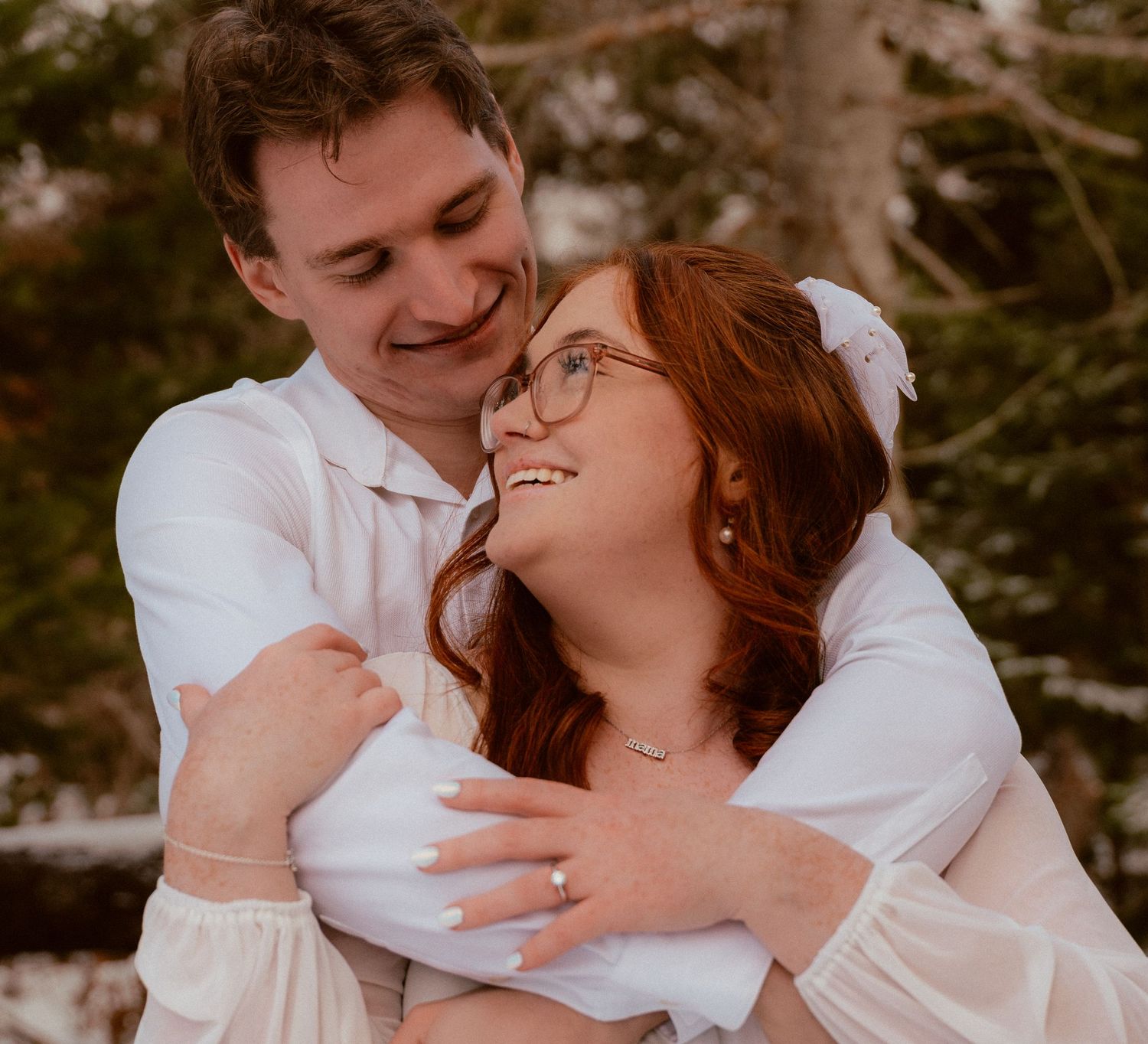A romantic couple embraces and smiles at each other while wearing white outfits against an outdoor backdrop.