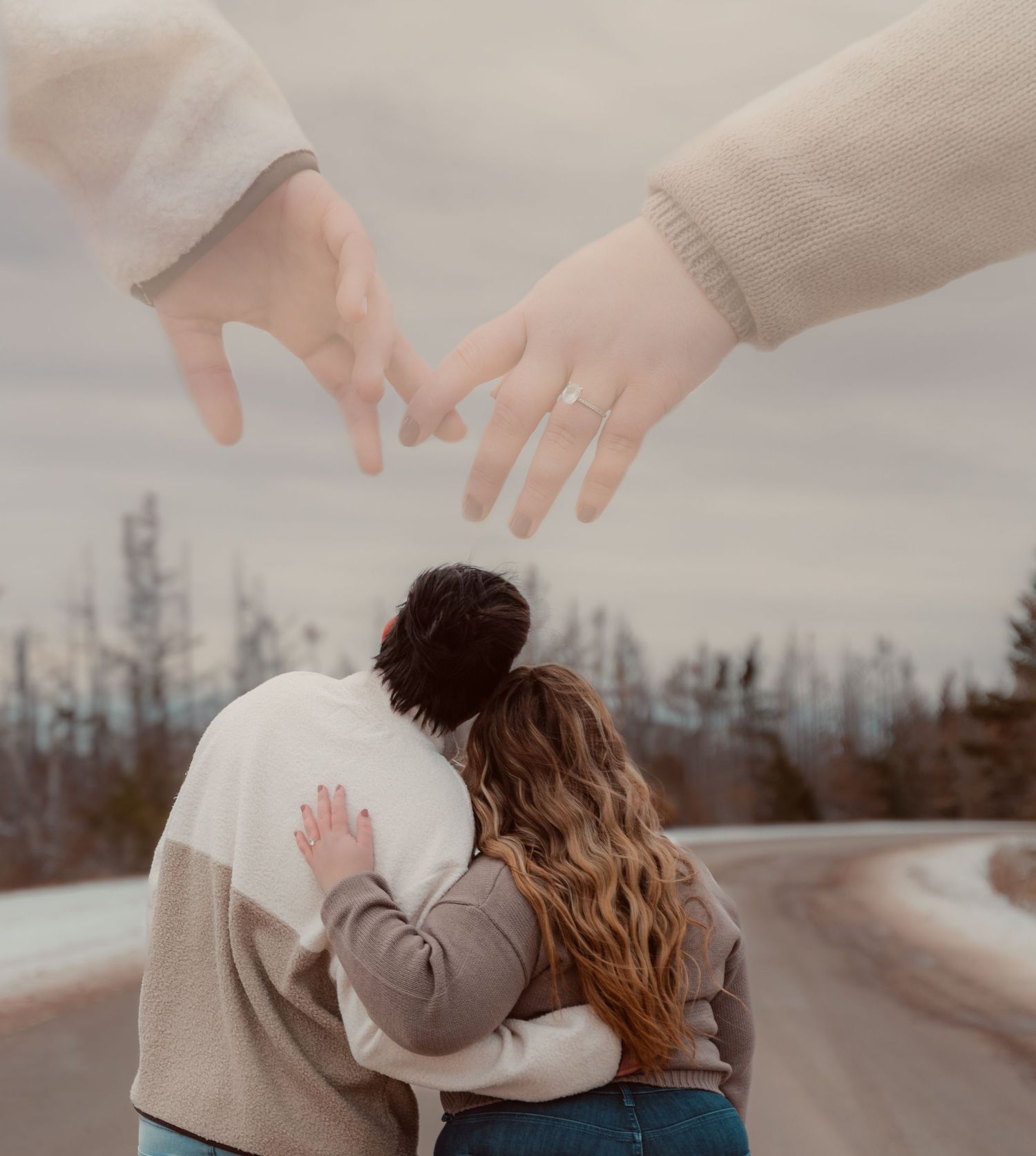 Two people embracing on a winding road with mountains in background as hands reach to create heart shape above.