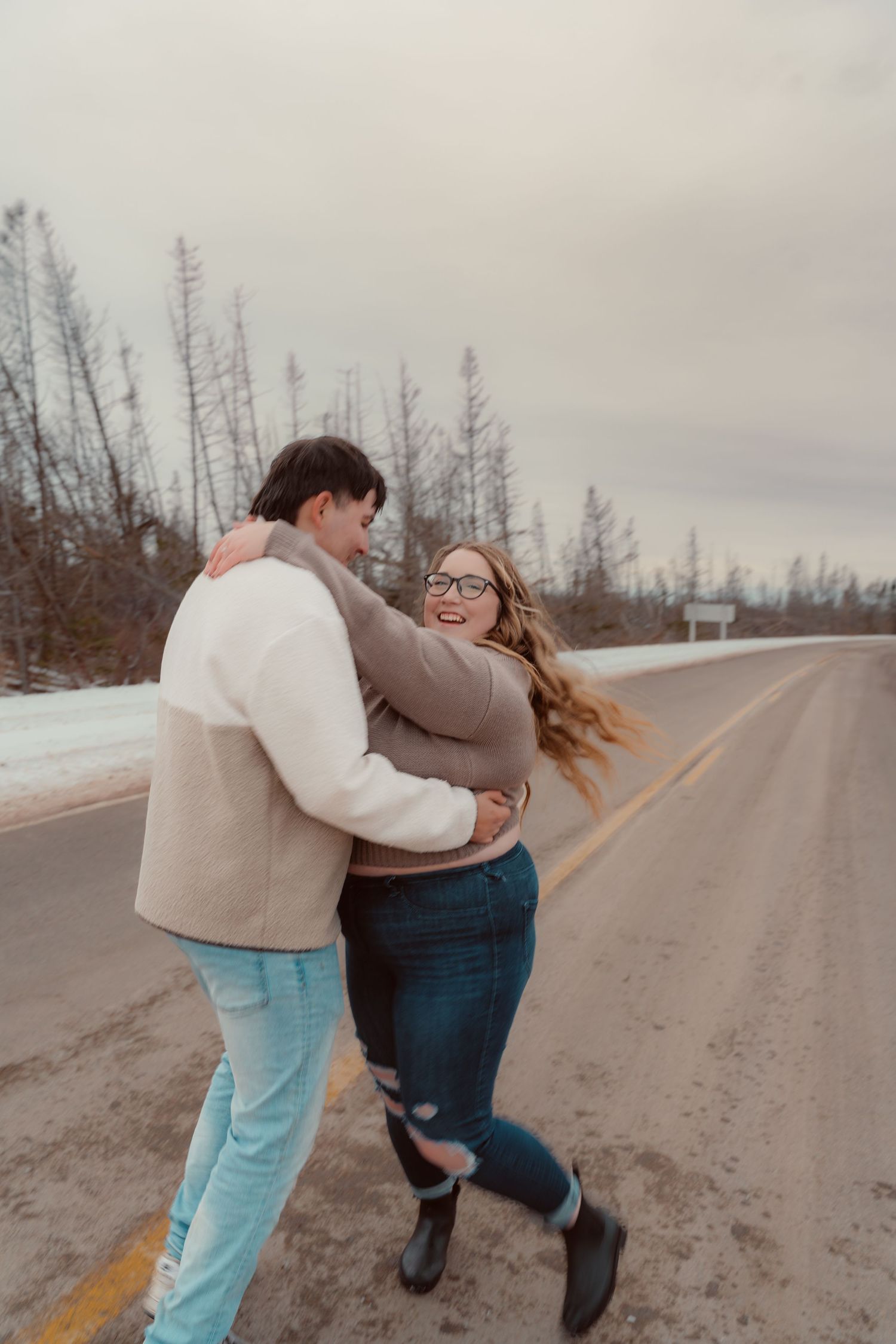 A couple embraces and dances playfully on an empty winter road with bare trees in the background.