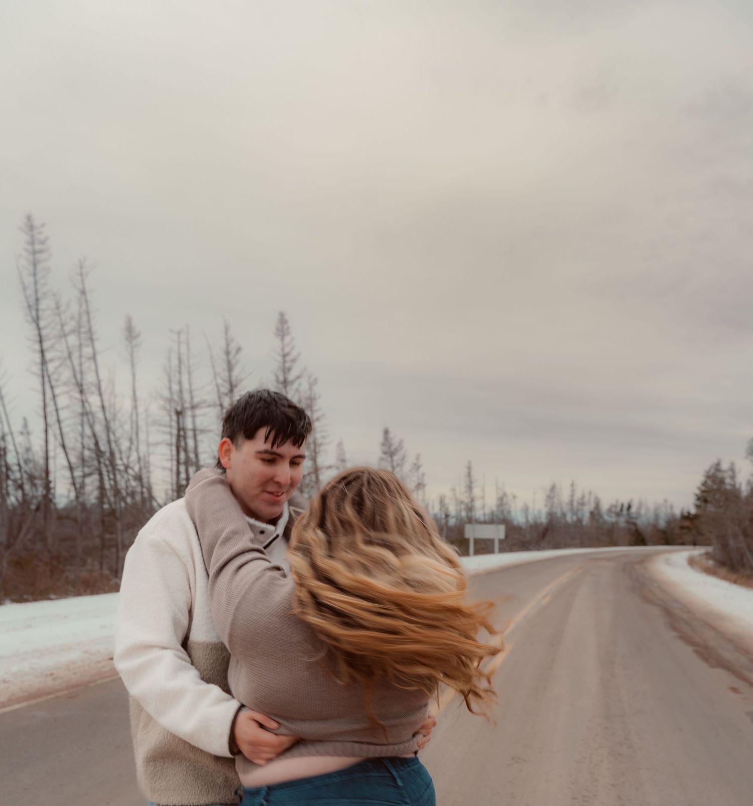 A couple embraces on a snowy winter road with barren trees in the background.
