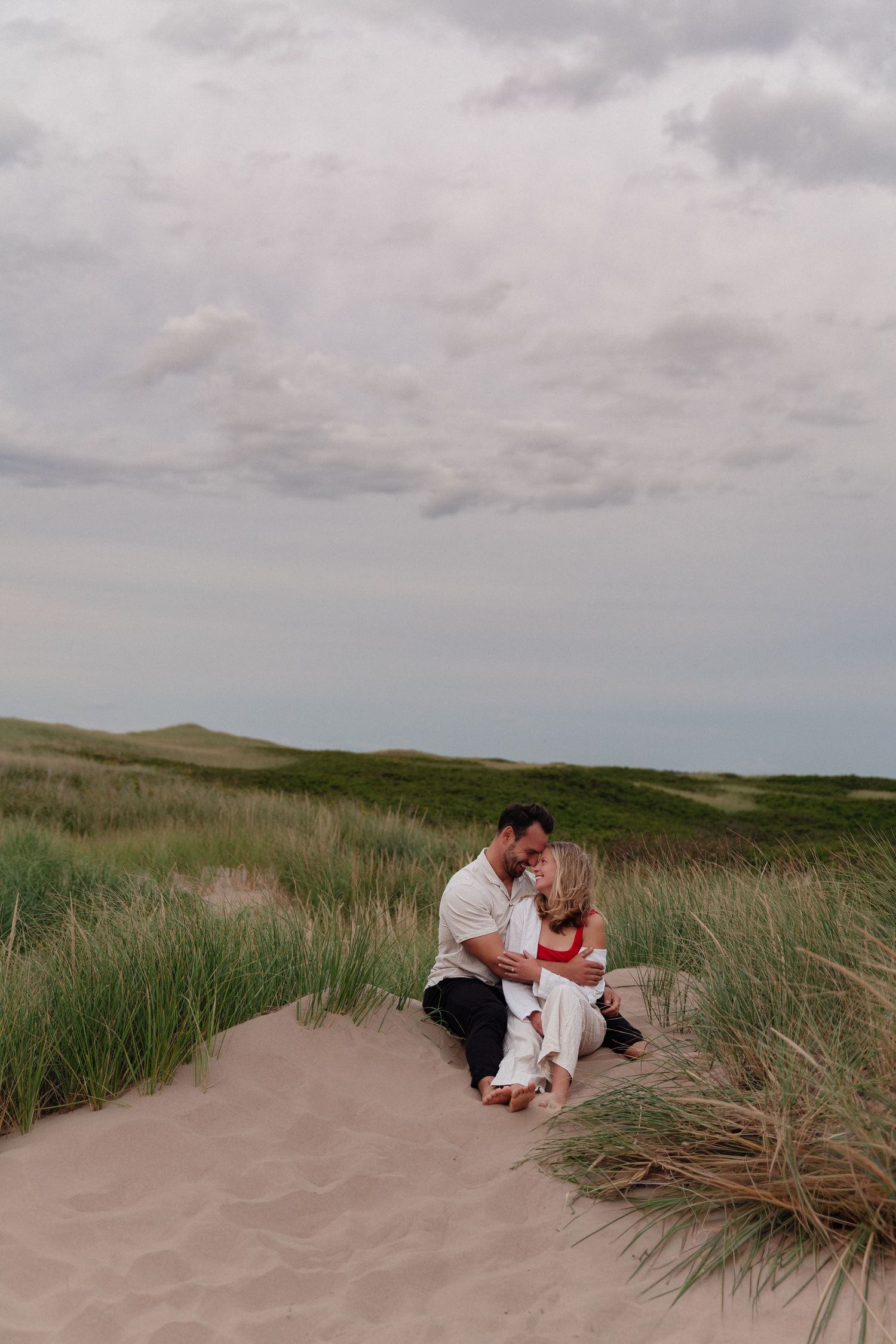 Couple sitting together on sand dunes surrounded by tall beach grass under cloudy coastal skies.