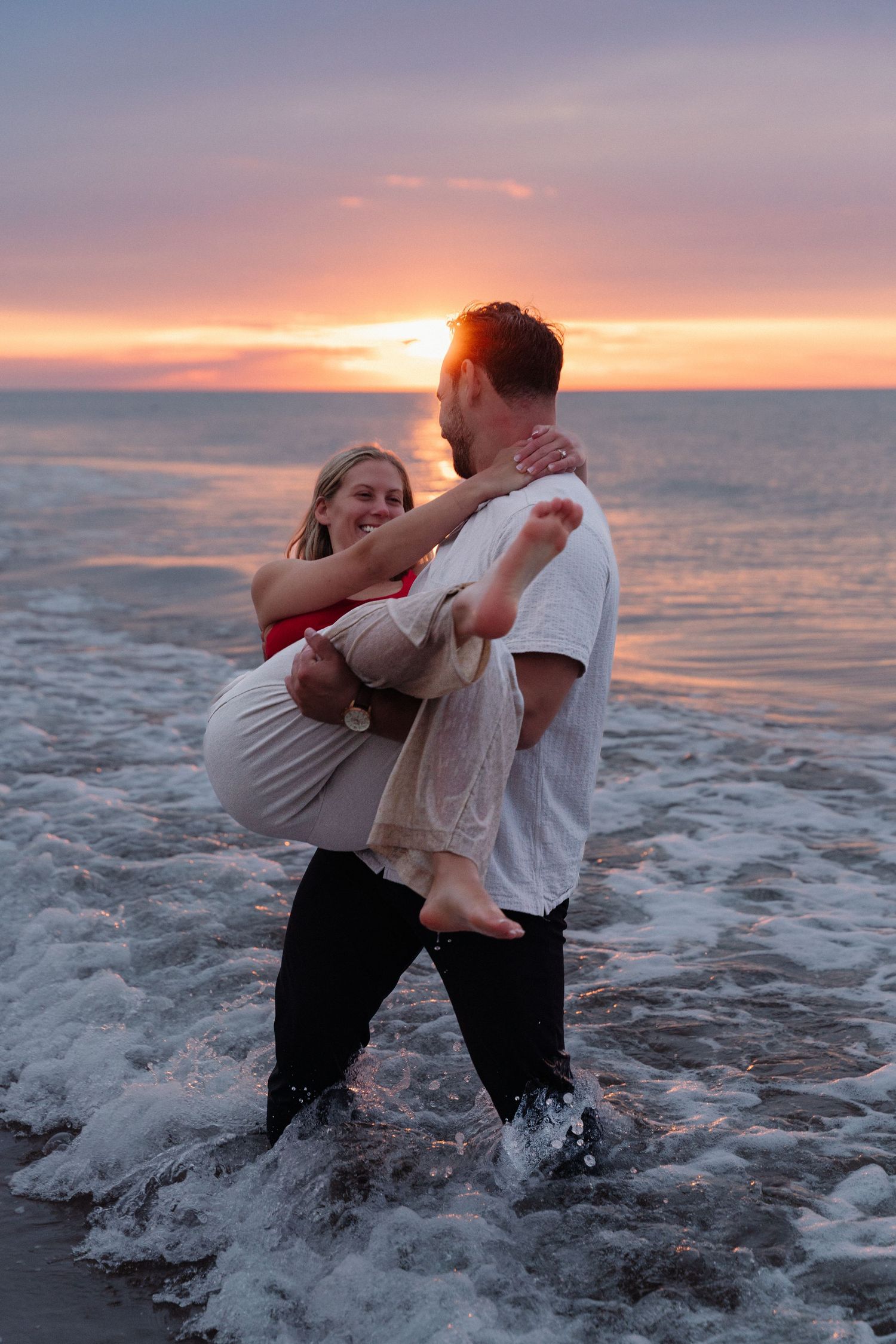 A romantic couple embraces while standing in ocean waves during a beautiful sunset on the beach.