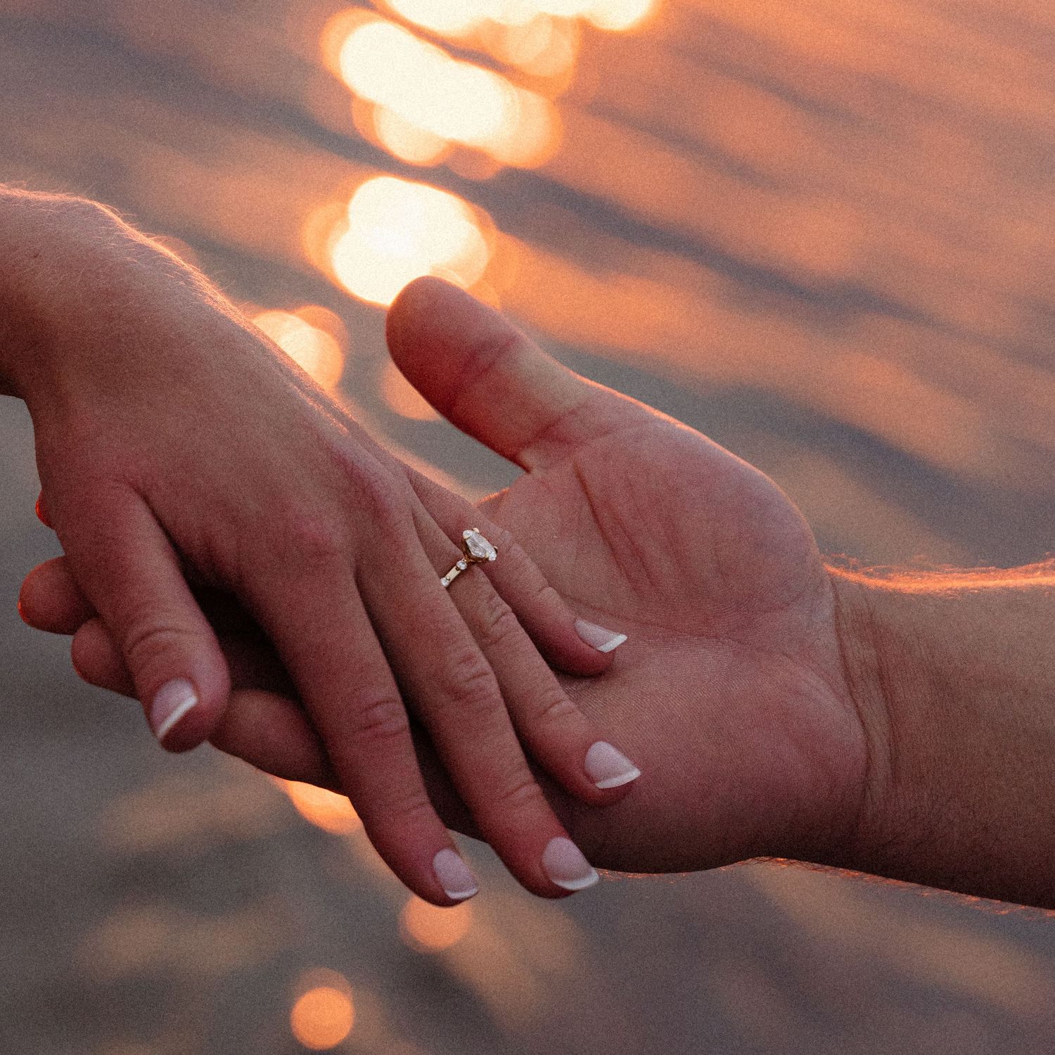 Close-up of hands holding each other against an orange sunset backdrop over water.