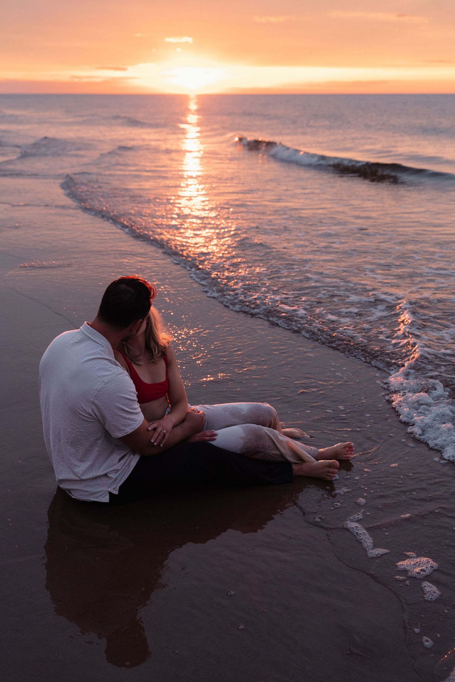 A couple embraces on a sandy beach at sunset as gentle waves roll onto the shore, creating a romantic silhouette.