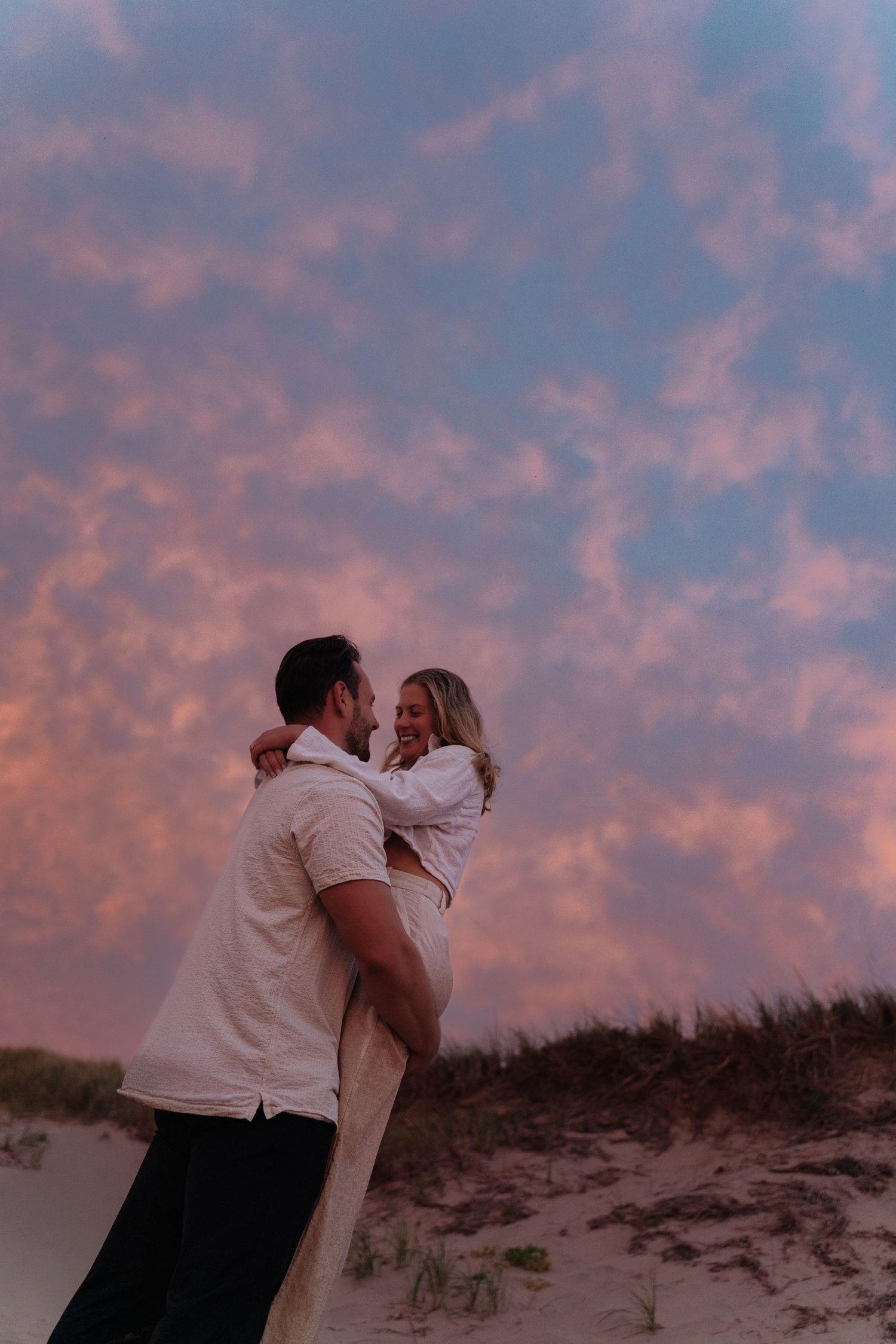 A couple shares an intimate moment against a stunning pink and purple sunset sky on sandy dunes.