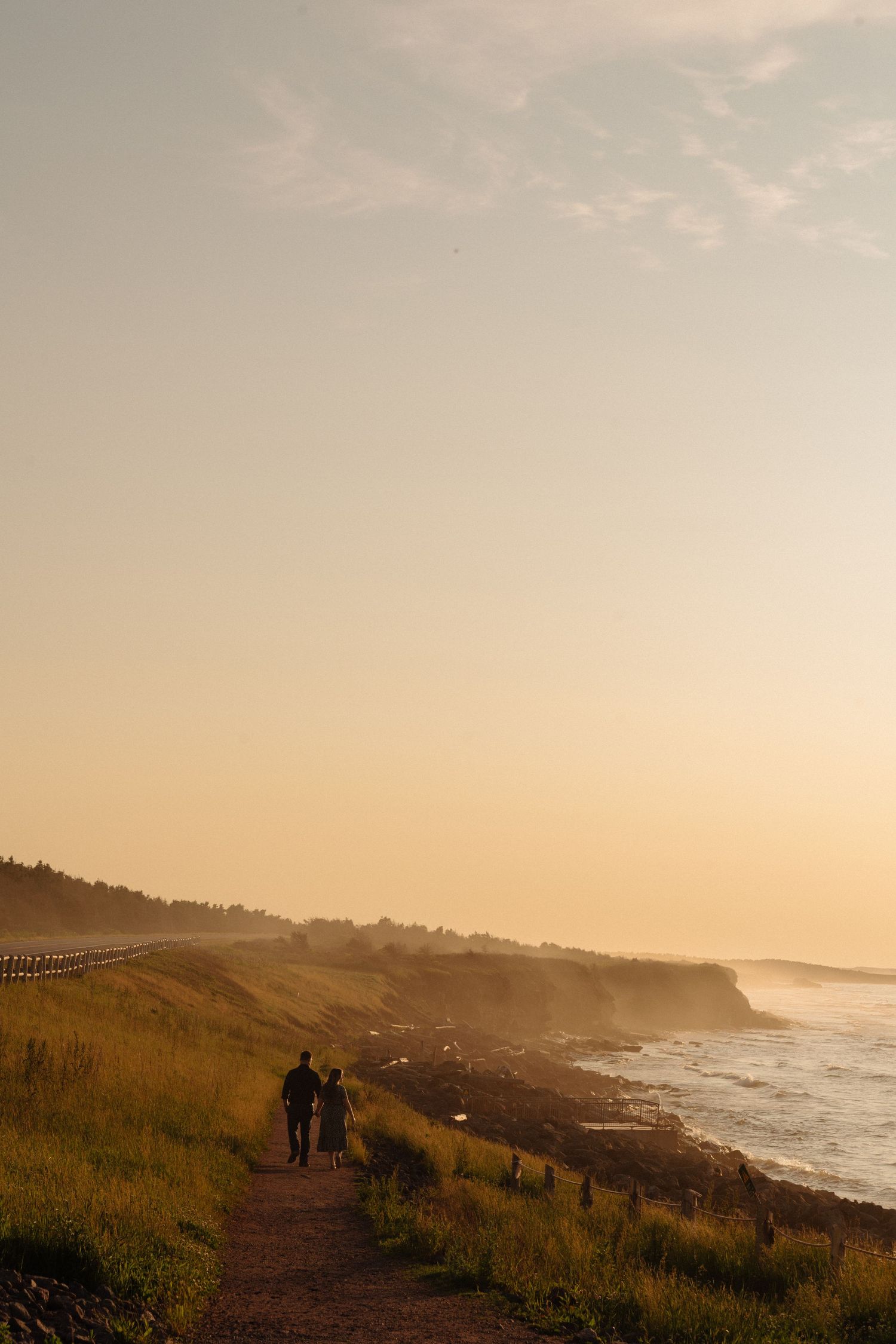 Silhouetted figures walk along a misty coastal trail at sunset with dramatic cliffs and crashing waves below.