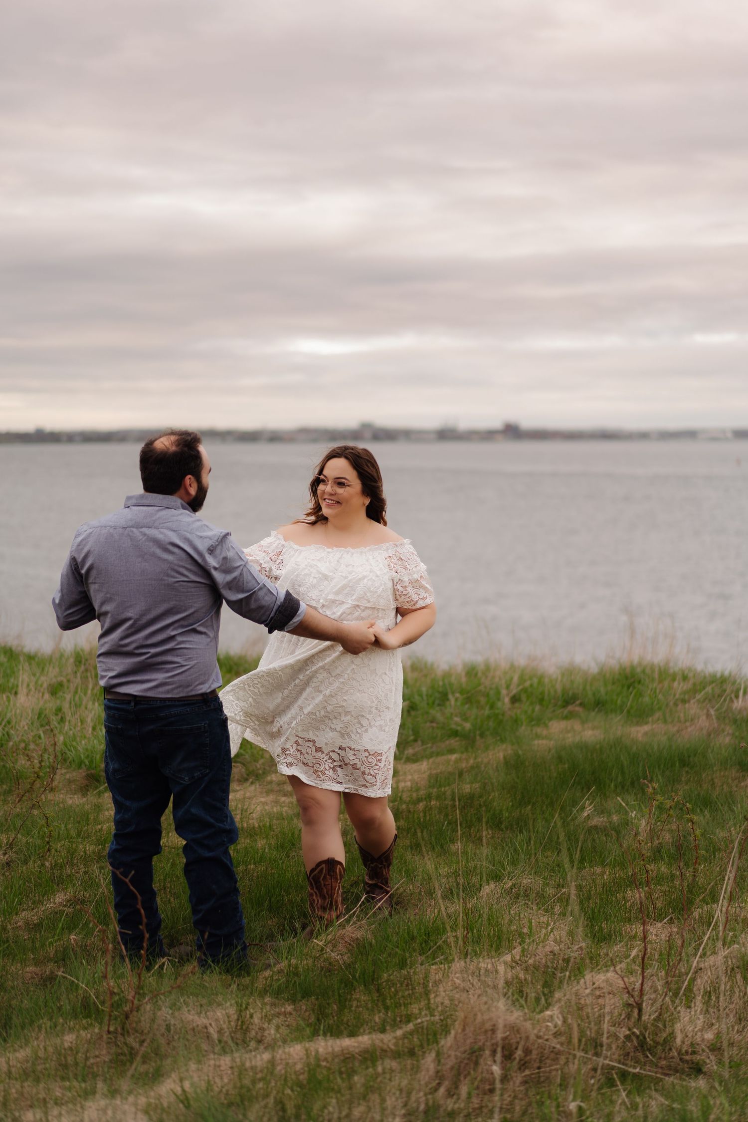 A couple in wedding attire poses together on a grassy hill overlooking a cloudy waterfront landscape.