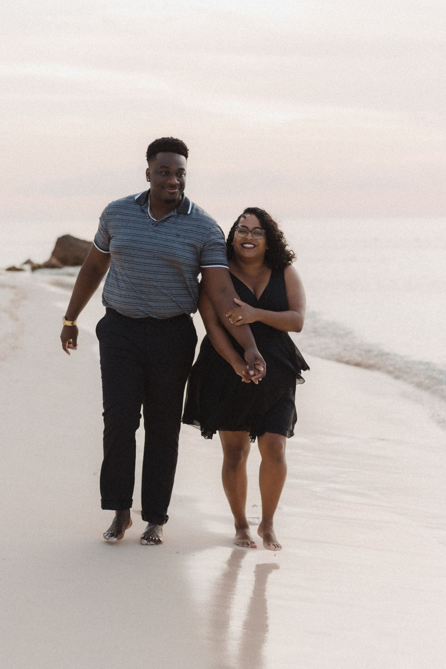 A couple walks barefoot together along a sunset beach, wearing casual attire and holding hands.