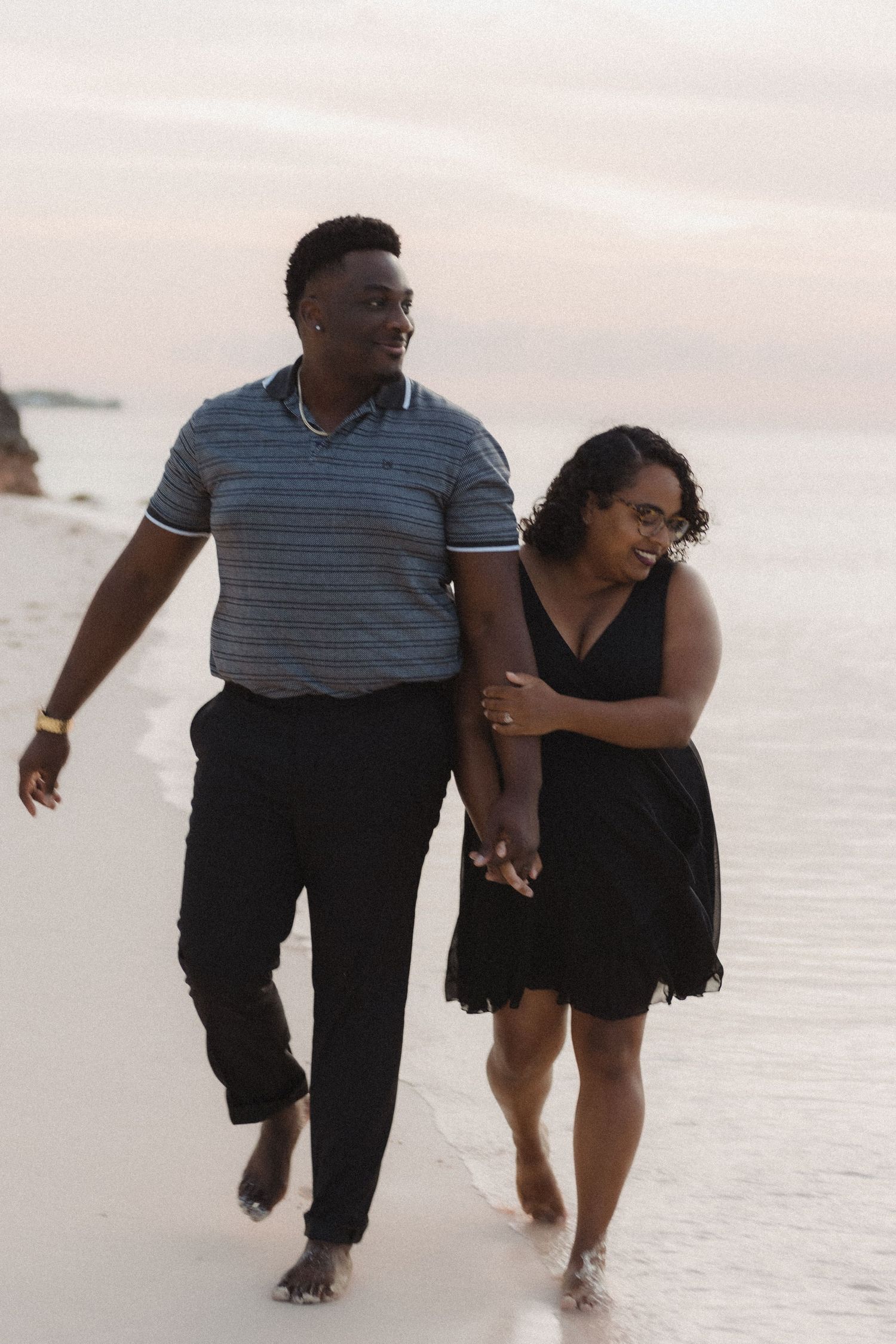 A couple walks hand in hand along the shoreline during a sunset beach photoshoot.