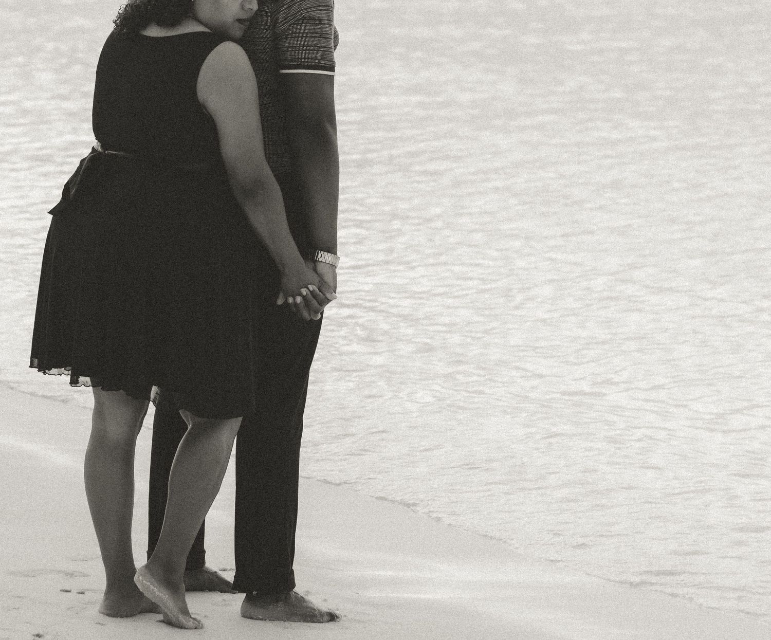 Black and white photo of a couple's legs as they stand together at the water's edge on a beach.