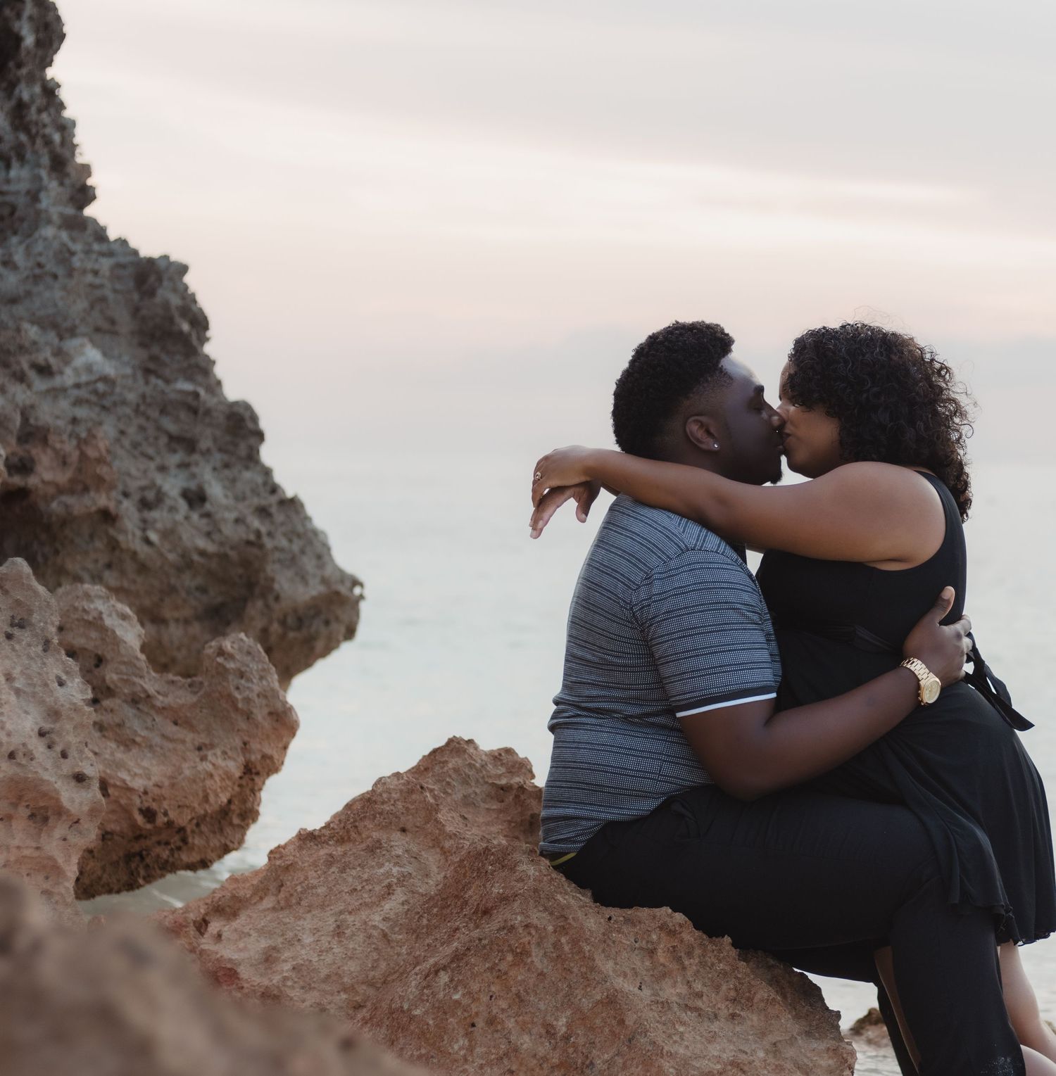 A couple embraces while sitting on rocky coastal cliffs overlooking the ocean at sunset.