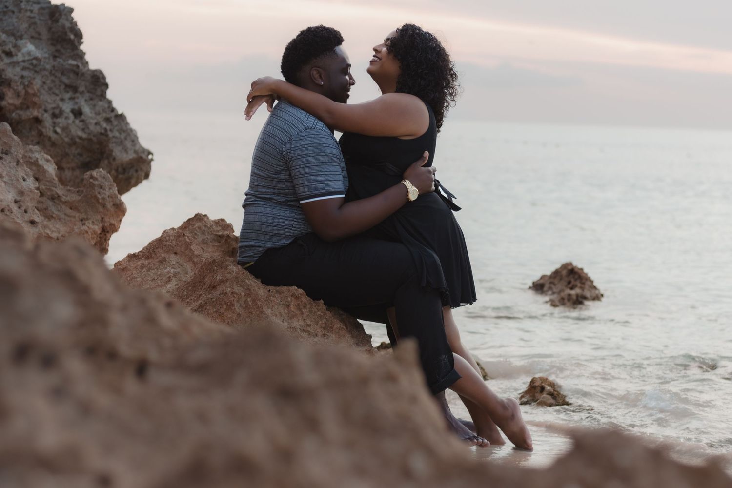 A romantic couple sits together on coastal rocks watching the sunset over the ocean.