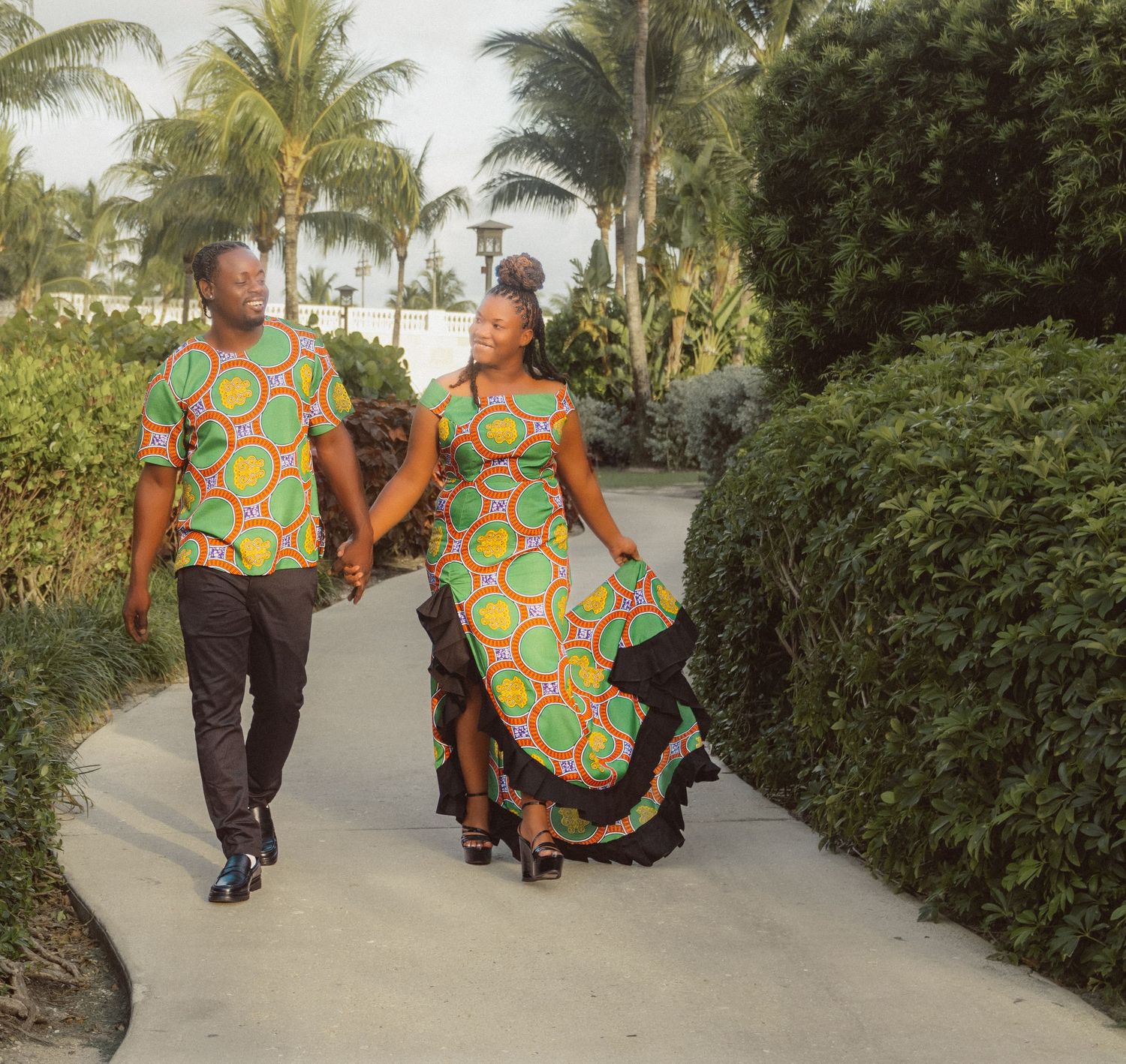 Couple in matching African print outfits walk along palm-lined pathway.